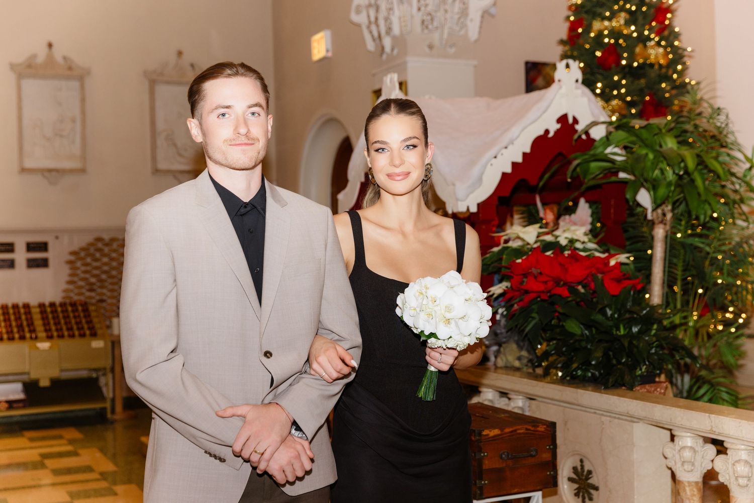 Elegant couple posing in decorated church holding white bouquet during Christmas wedding celebration.