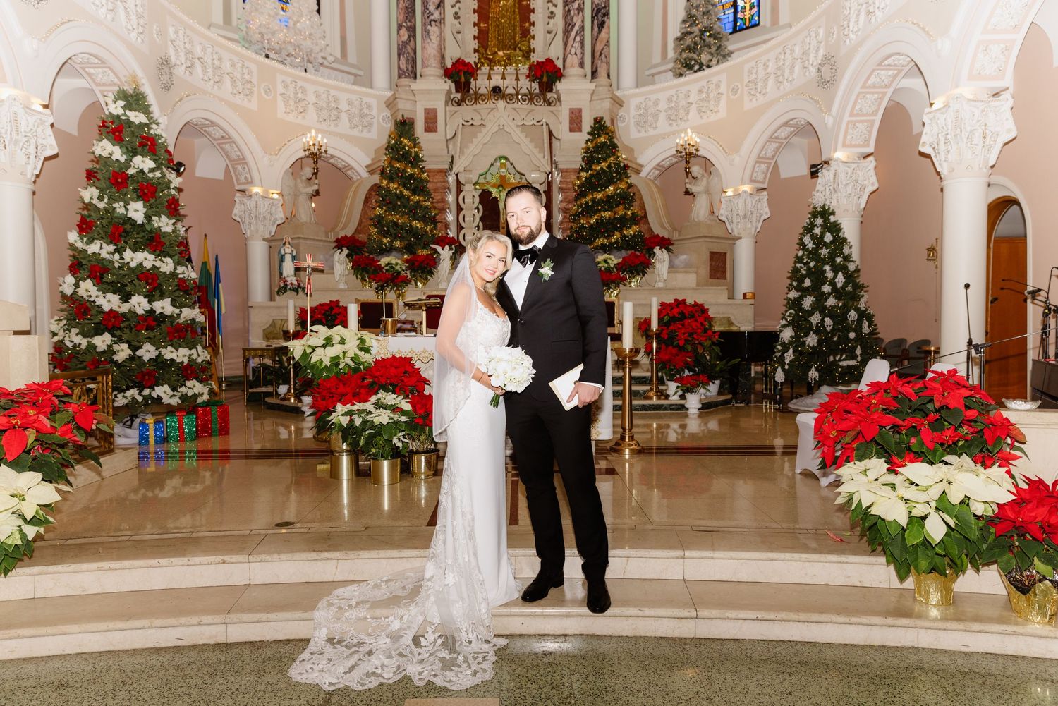 Newlyweds stand together in ornate church aisle beautifully decorated with Christmas trees and red and white poinsettias.