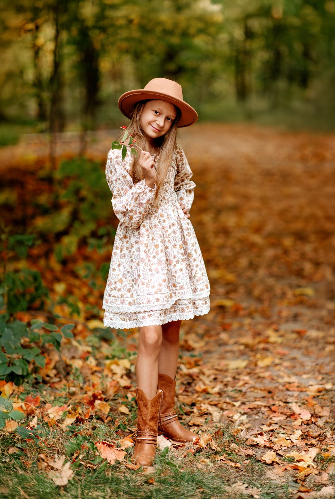 Young girl in floral dress, tan cowboy hat and brown boots poses on autumn leaf-covered path in forest.