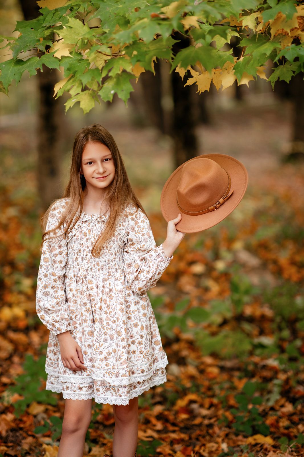 Young girl in white floral dress holding tan wide-brim hat under yellow maple tree in autumn park.