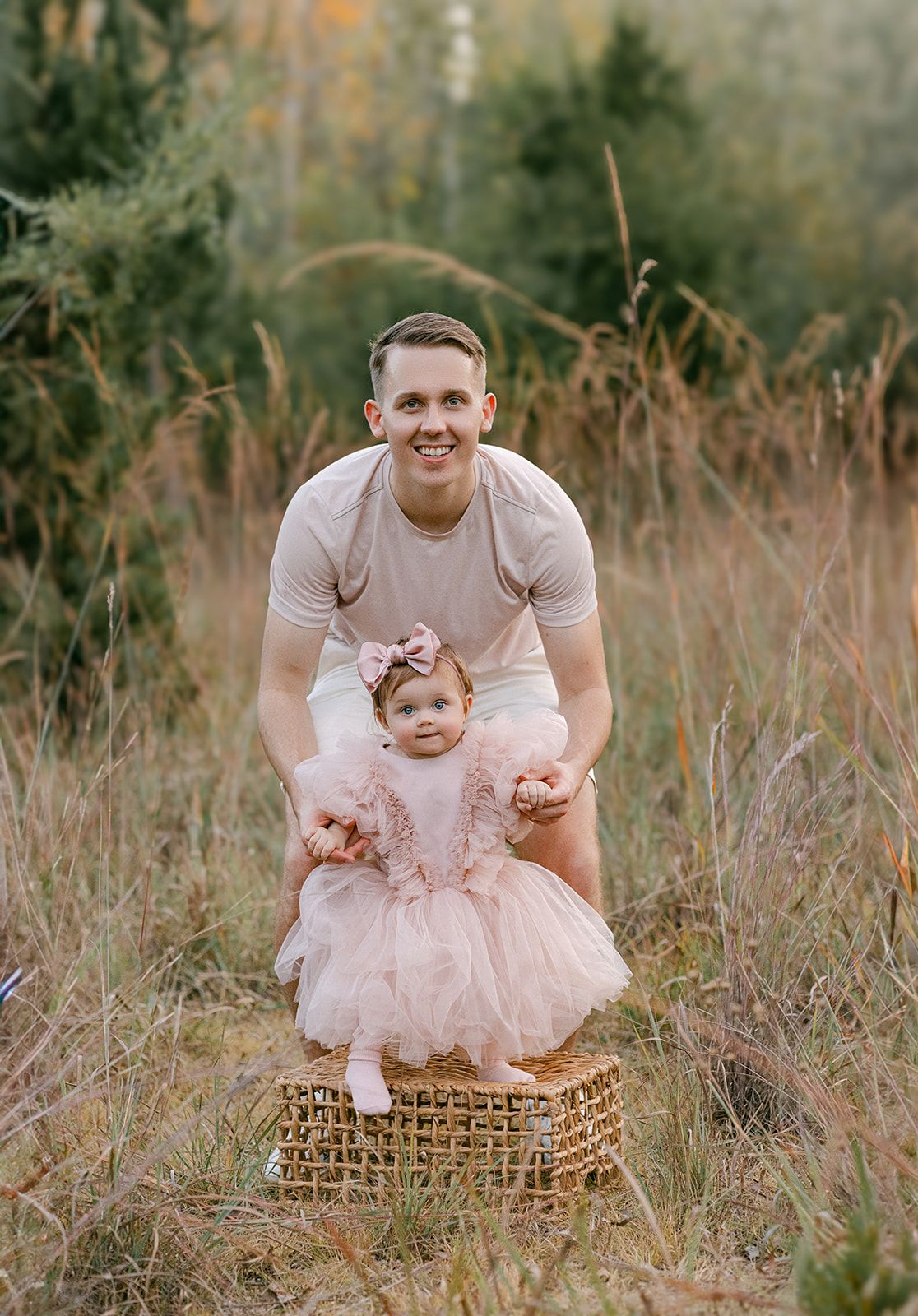 Smiling father poses with baby girl in pink tutu dress standing in wicker basket outdoors.