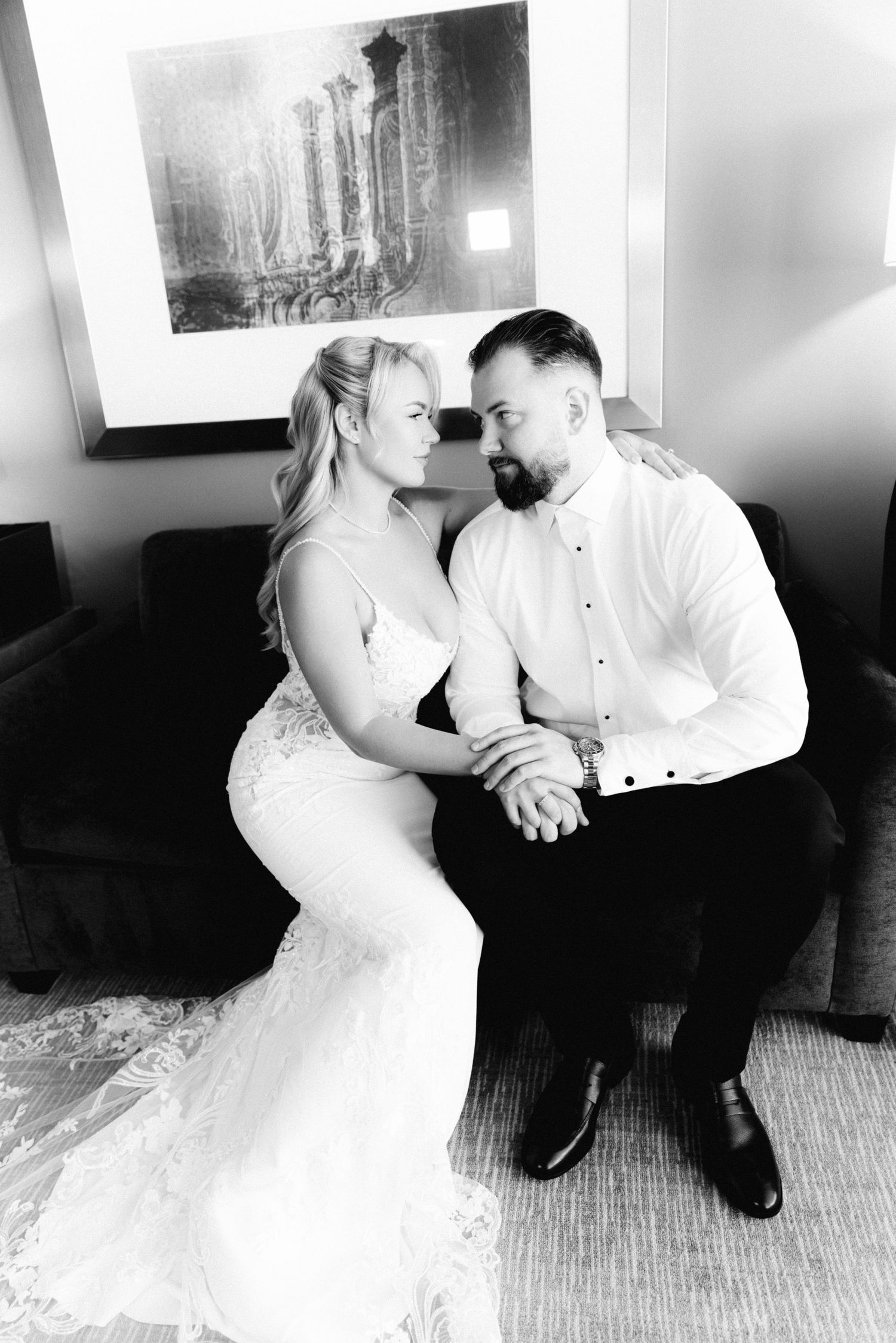 Black and white photo of bride and groom sitting close together, holding hands and gazing at each other.