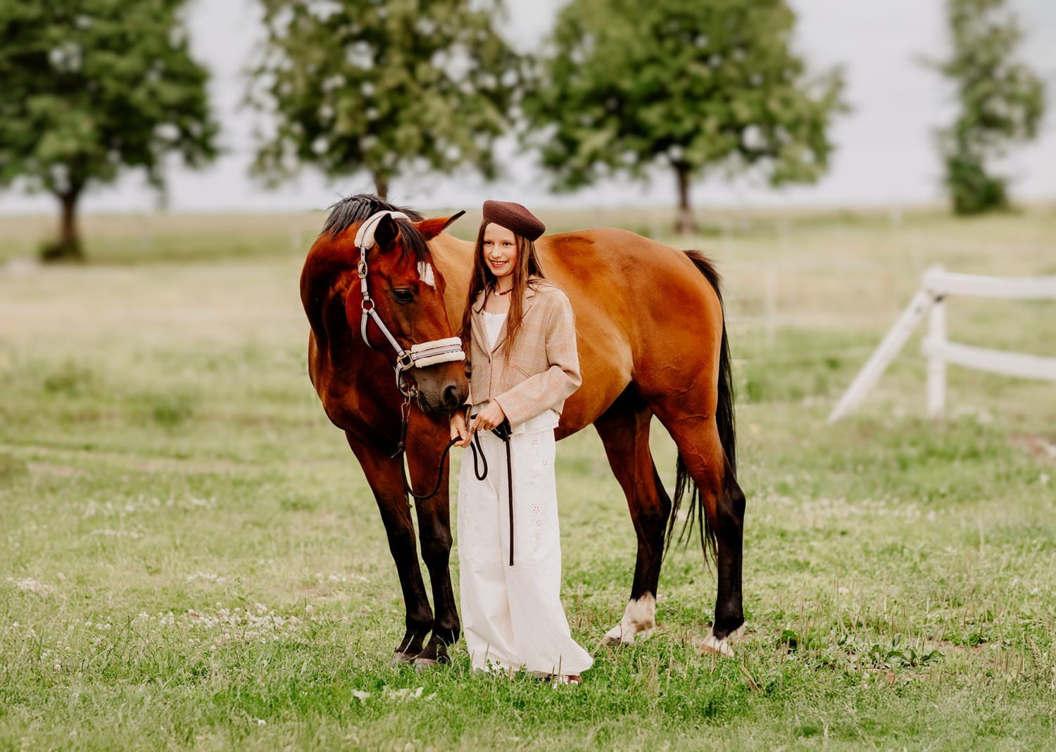 Young woman in white dress standing beside a chestnut horse in a green field with trees in background.