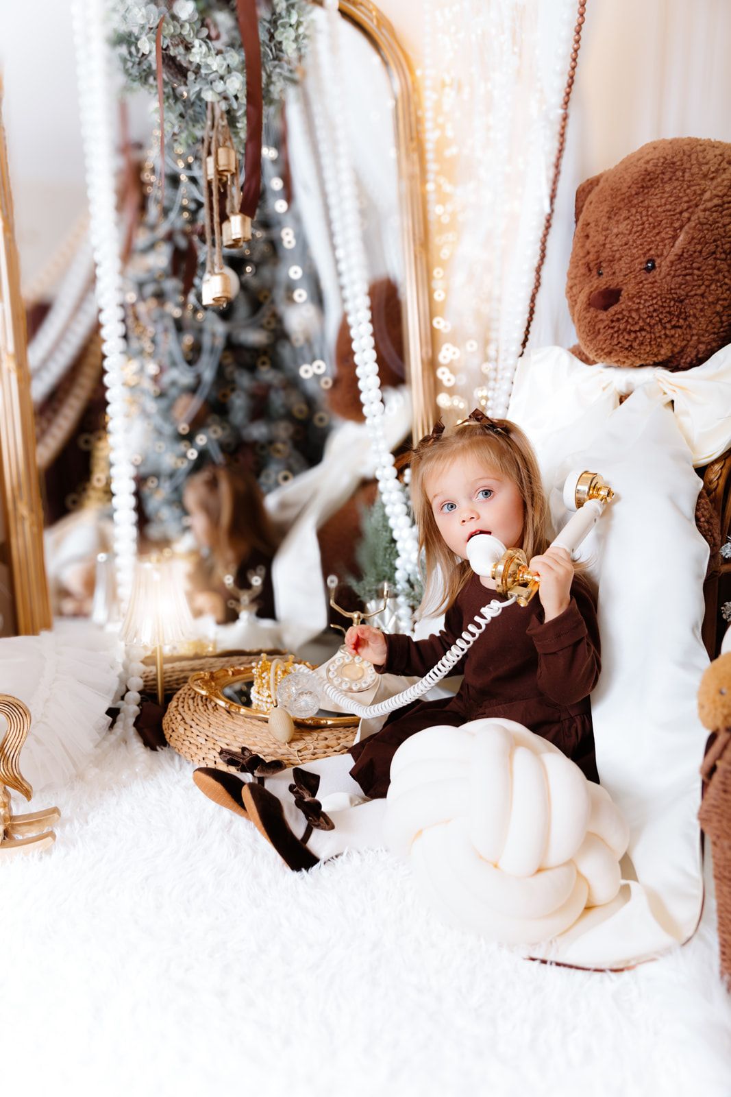 Young girl in brown dress sits by Christmas tree with giant teddy bear, holding golden scepter on white fur rug.