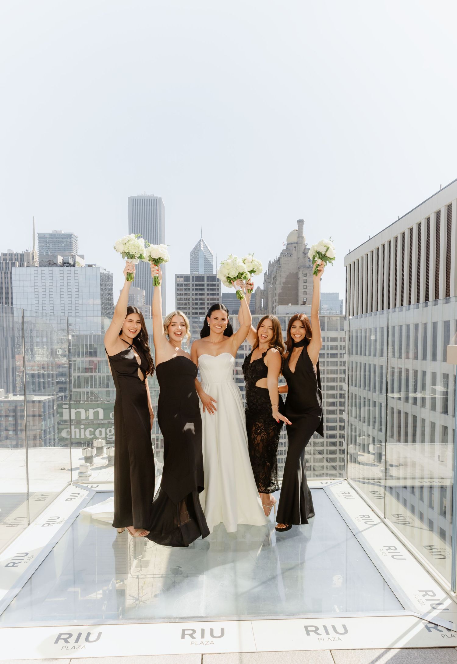 Bride and bridesmaids in black dresses raise bouquets on a rooftop at Hotel RIU with Chicago skyline backdrop.