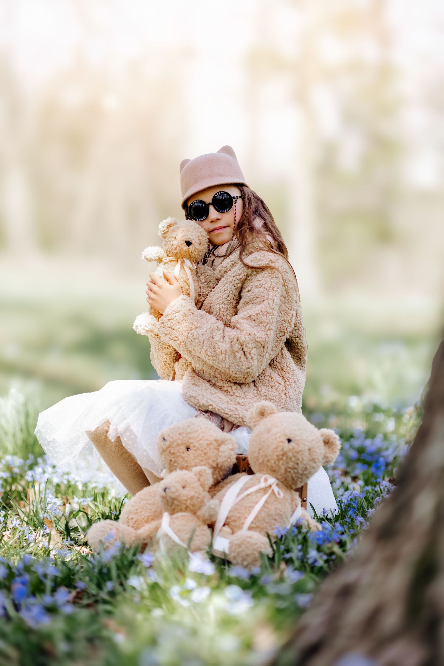 Young girl in beige fur coat and sunglasses hugging teddy bear in spring flower field.