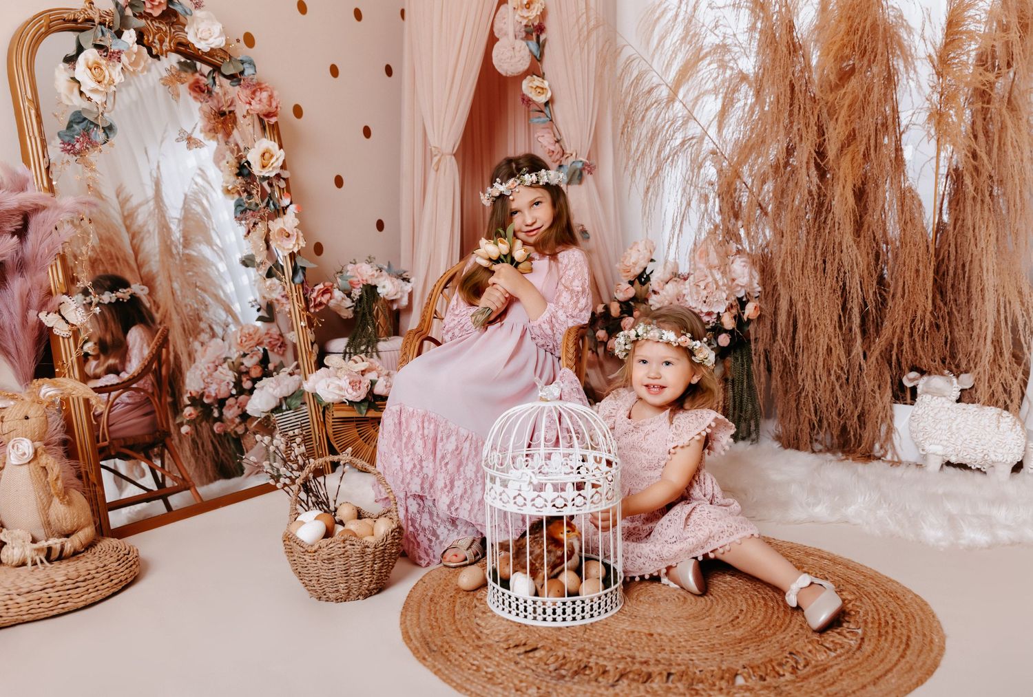 Two young girls in pink dresses and floral crowns pose in a boho floral studio with pampas grass, a birdcage, and mirror.