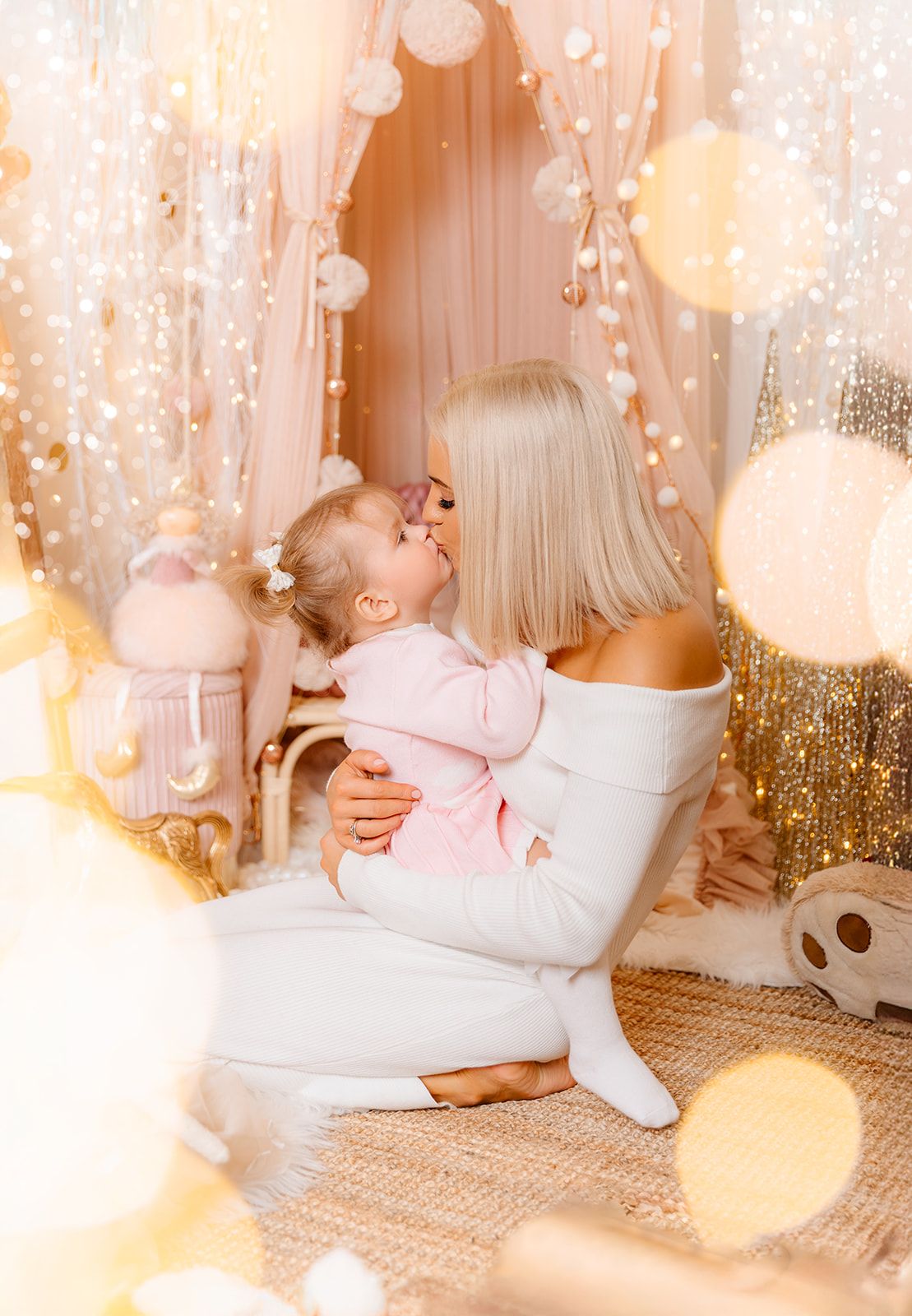 Blonde mother in white dress kissing baby girl in pink outfit in dreamy bokeh light decorated room.