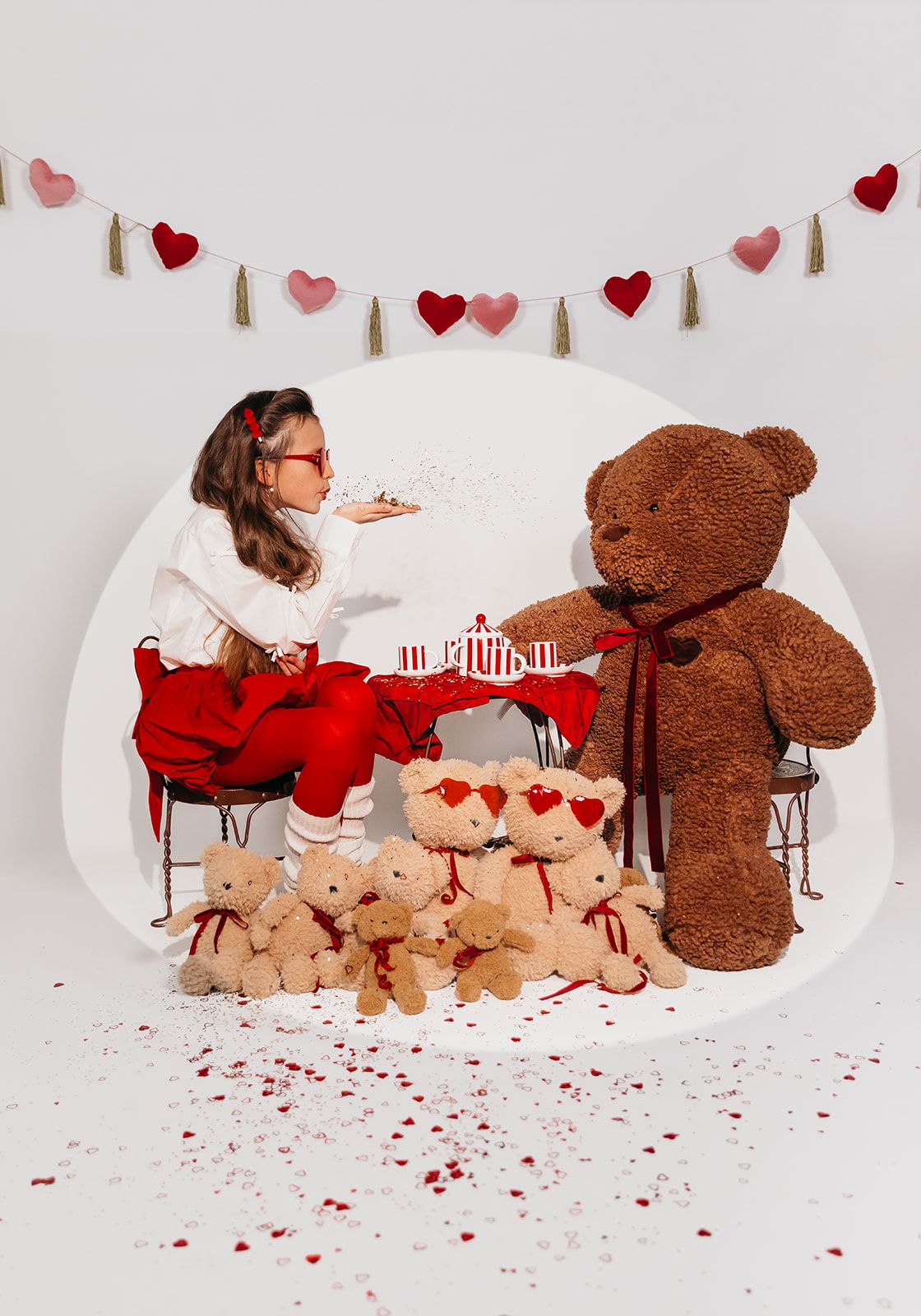 Girl having a Valentine's Day tea party with large and small teddy bears, heart garland backdrop, red confetti floor.