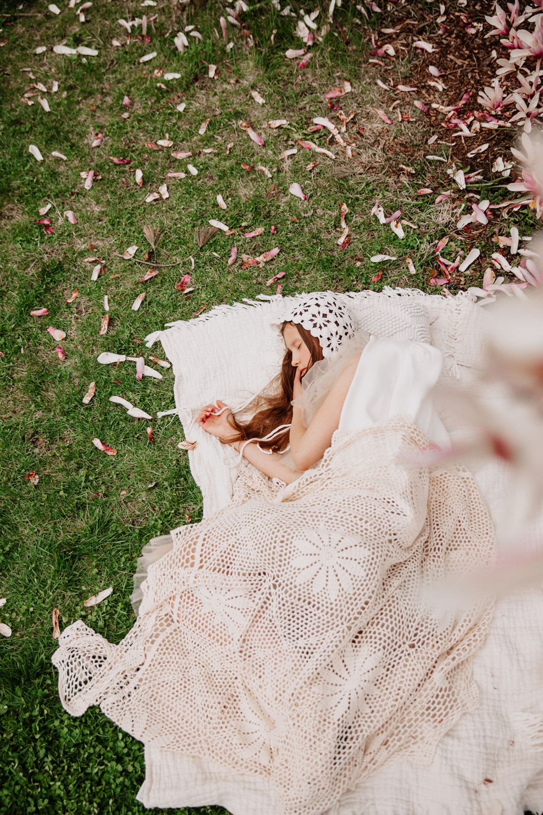 Aerial view of a woman lying on a white crochet blanket on green grass with pink magnolia petals falling around her.