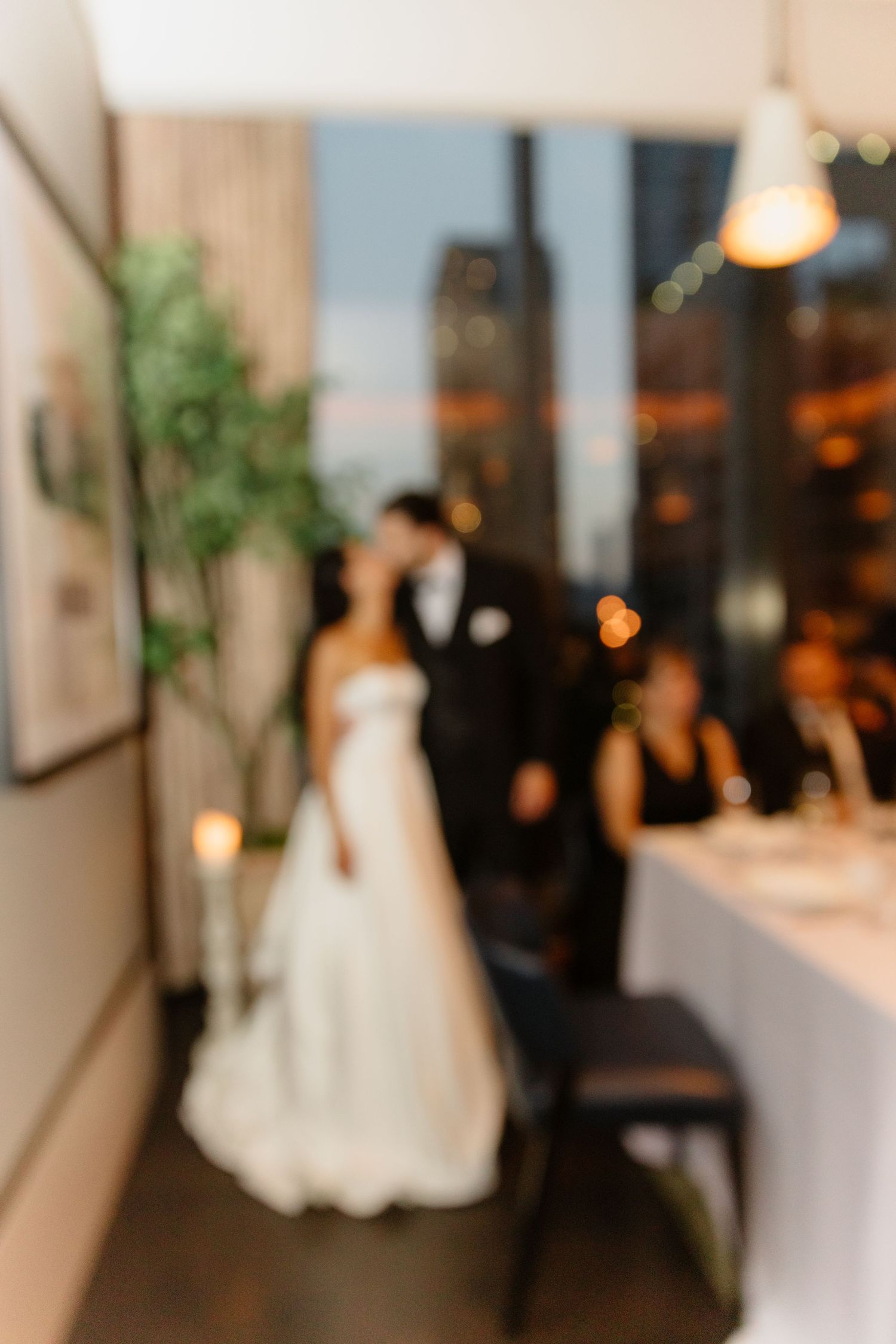 Blurred romantic image of bride and groom sharing a kiss by large windows at evening wedding reception.