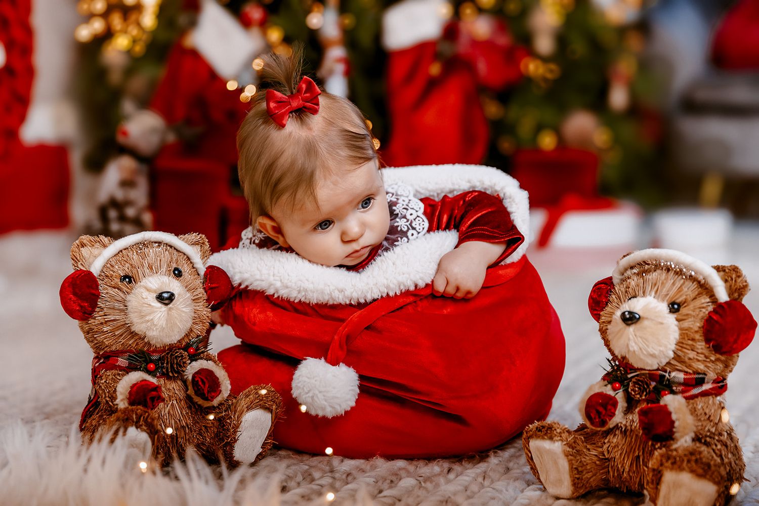 Baby in Santa outfit sitting in red bag with teddy bears wearing reindeer costumes near Christmas tree.