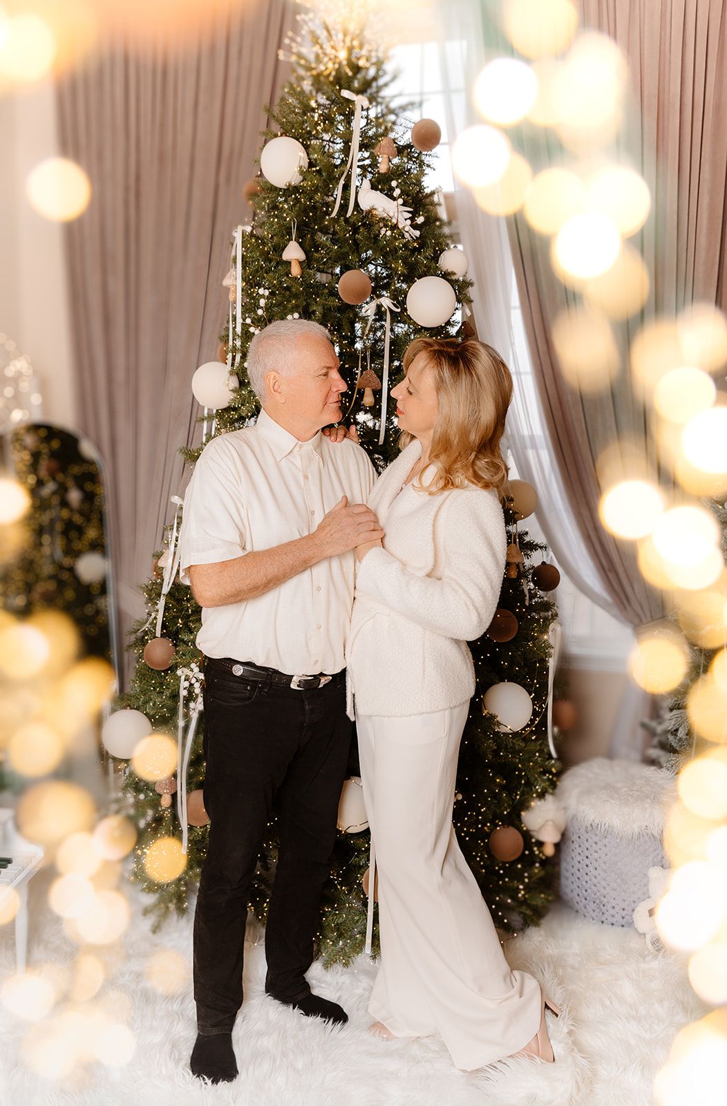 Couple in white outfits embracing near decorated Christmas tree with bokeh lights.