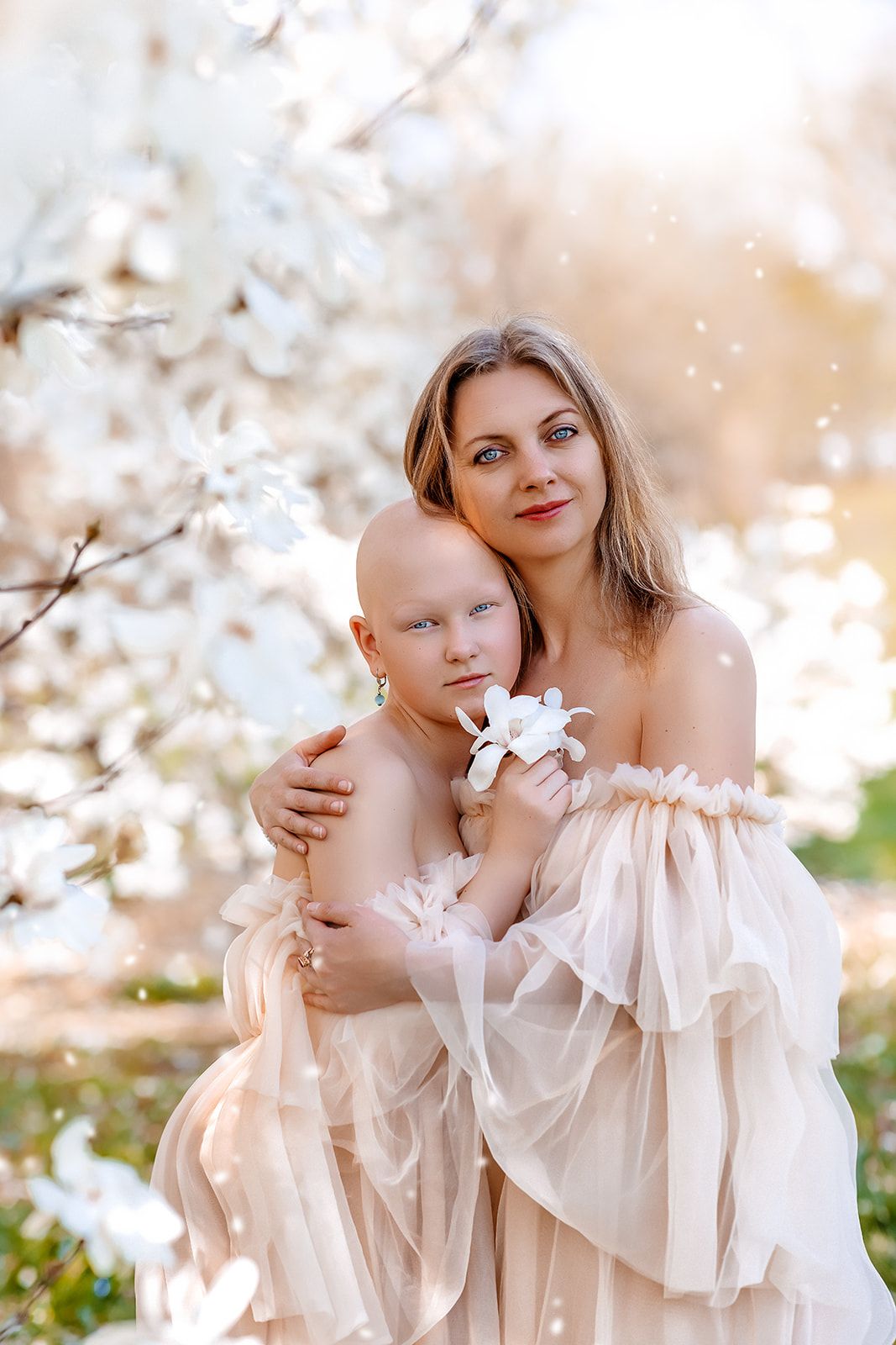 A mother in a blush gown embraces her bald child holding a white flower among blooming magnolia trees.
