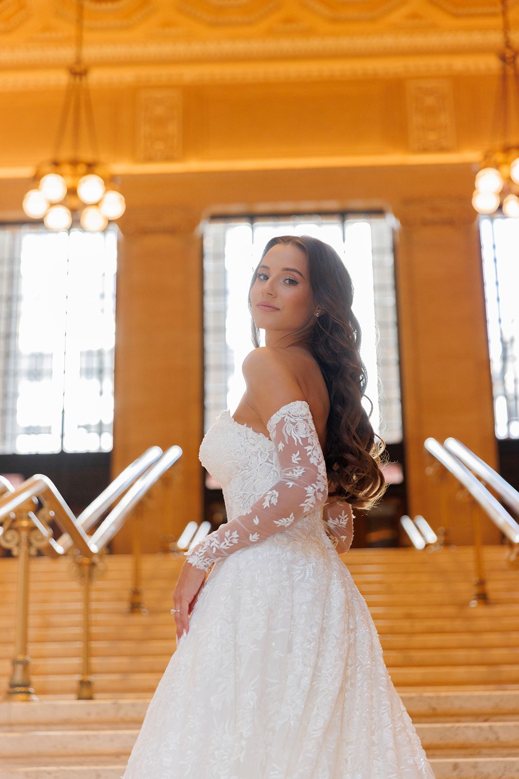 Bride in lace off-shoulder gown poses on grand staircase in elegant golden hall.