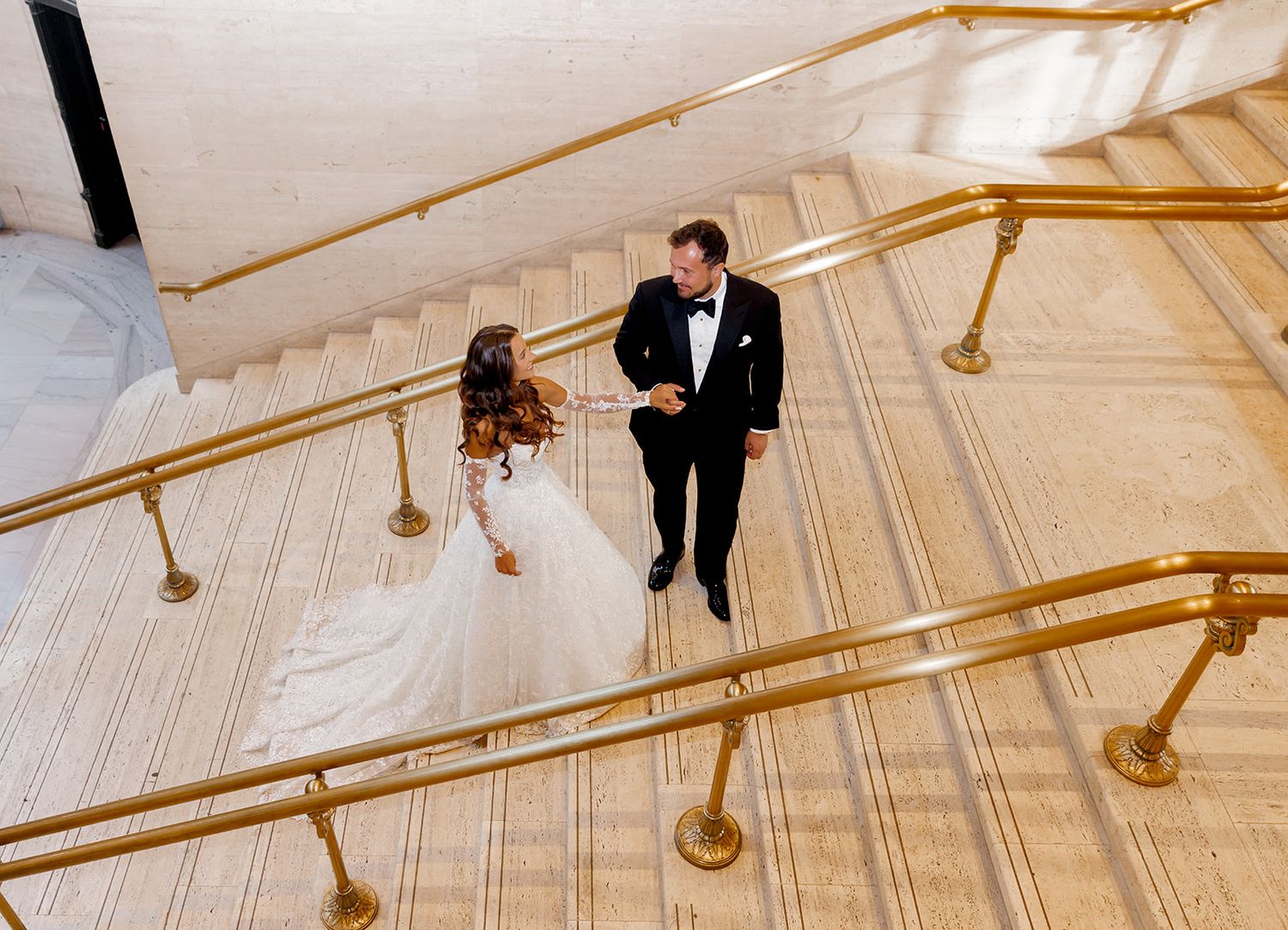 Bride and groom share a romantic moment on elegant marble stairs with gold railings at a wedding venue.
