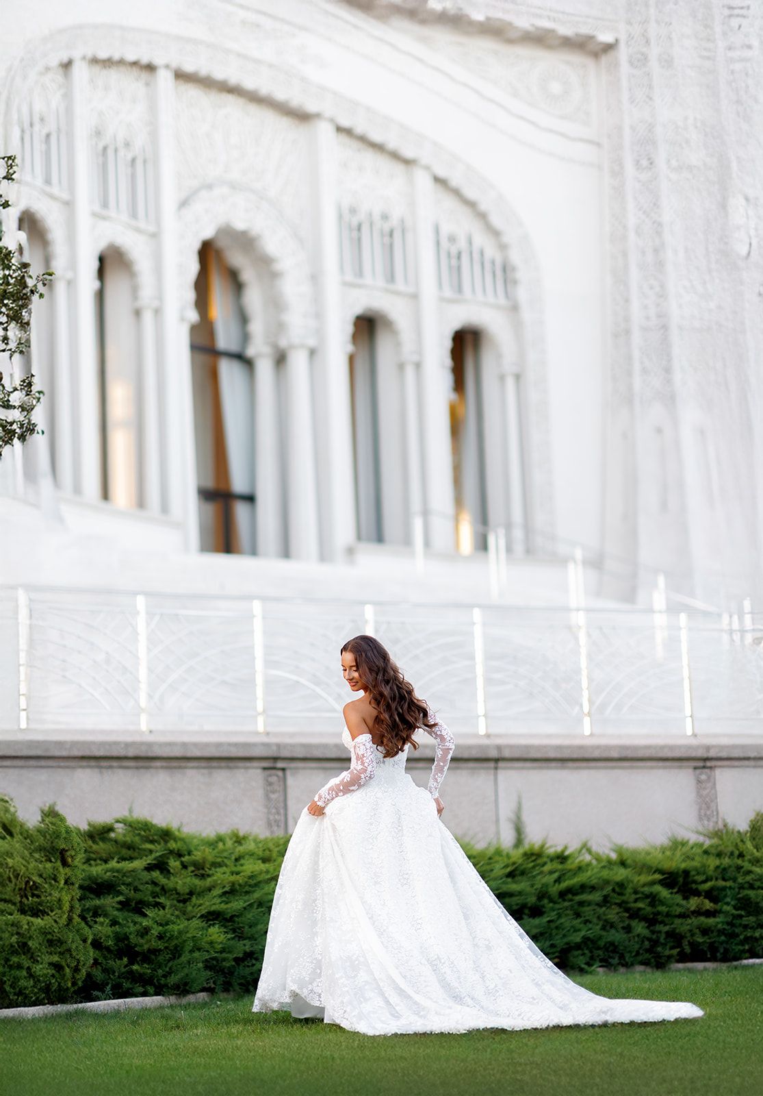 Bride in lace long-sleeve wedding gown with cathedral train poses before ornate white building.