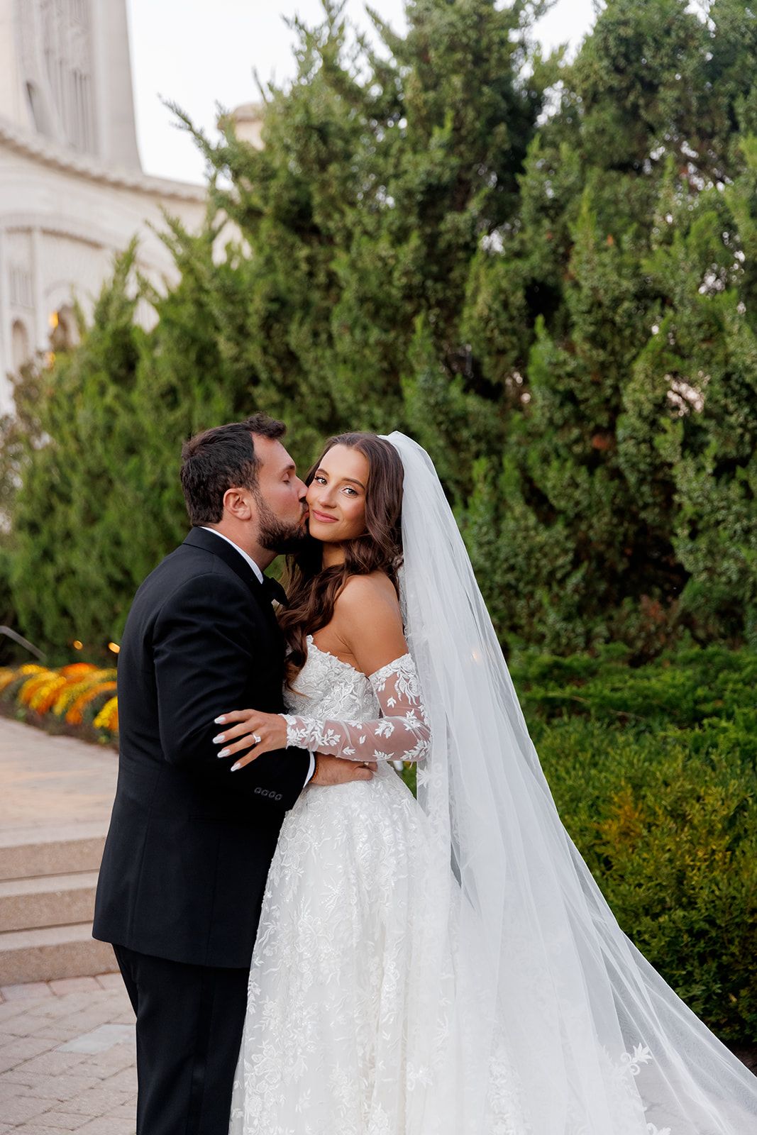 Groom kisses bride on the cheek outdoors near tall evergreen trees, bride smiling in lace wedding gown with long veil.