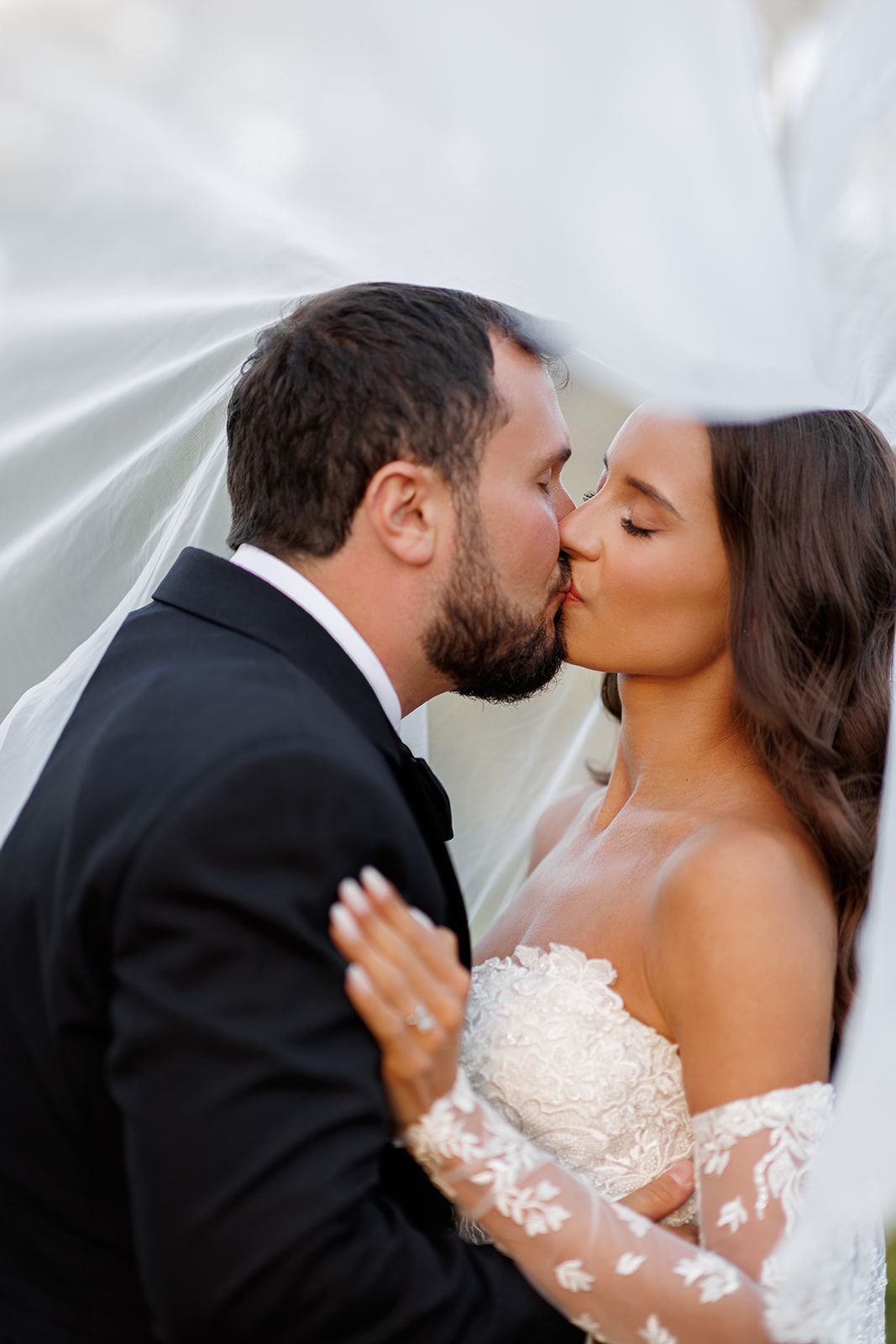 Bride and groom sharing a romantic kiss under a flowing white veil on their wedding day.