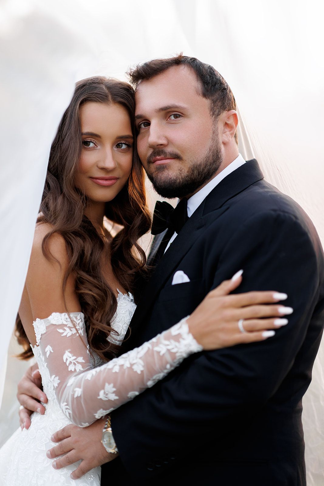 Elegant bride and groom pose closely together under a white veil, smiling at the camera.