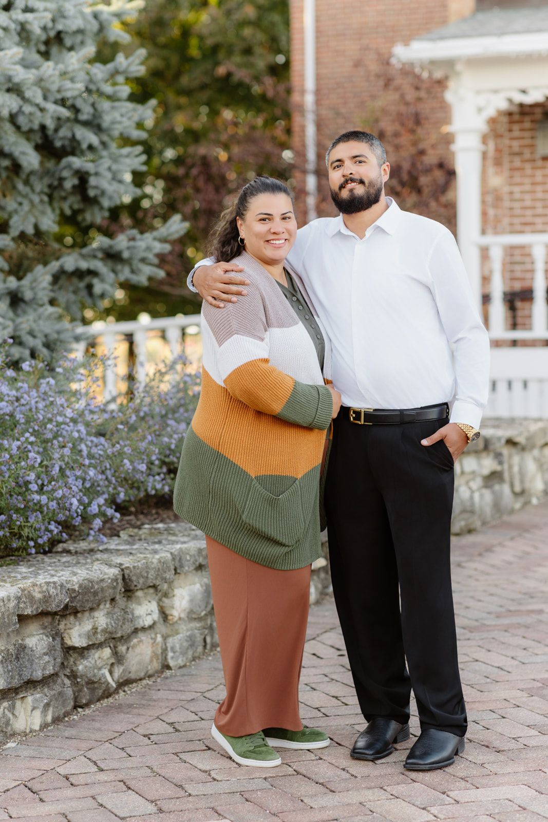 Smiling couple embraces outdoors on a brick pathway surrounded by lush greenery and purple flowers.