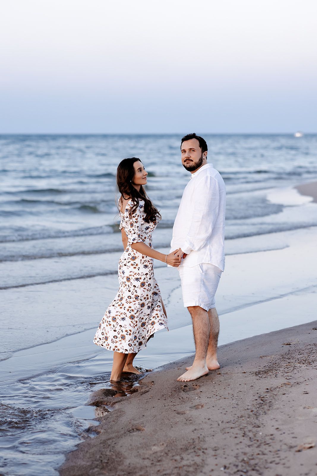 Couple holding hands walking barefoot on a sandy beach at dusk, woman in floral dress, man in white outfit.
