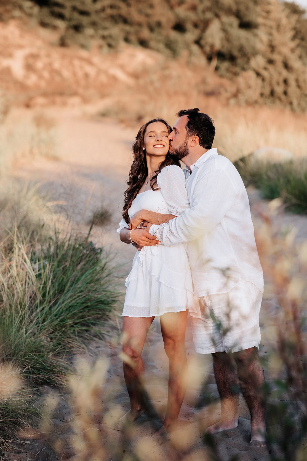 Man kissing woman on cheek while embracing her on sandy beach path at golden hour sunset.