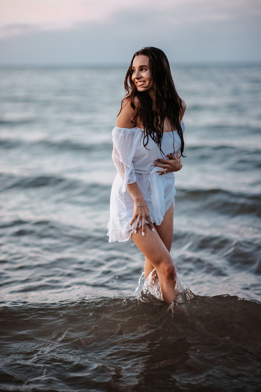 A smiling woman in a white off-shoulder dress stands in ocean waves at dusk, her dark hair wet and flowing.