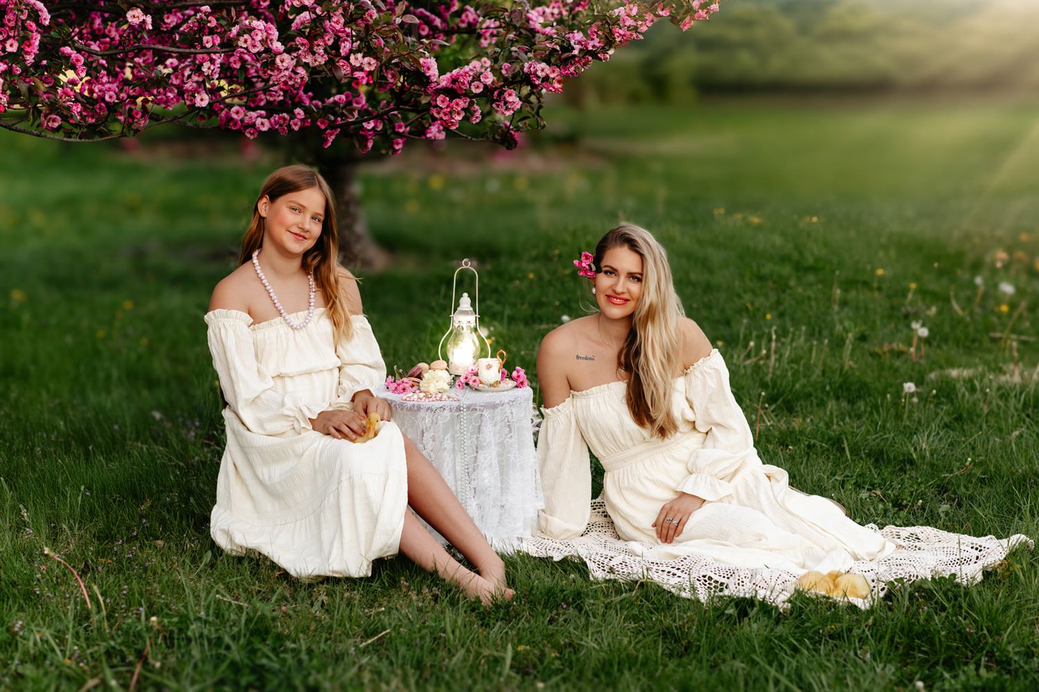 Two women in white dresses sit on grass under pink blossoming trees at a decorative outdoor picnic.