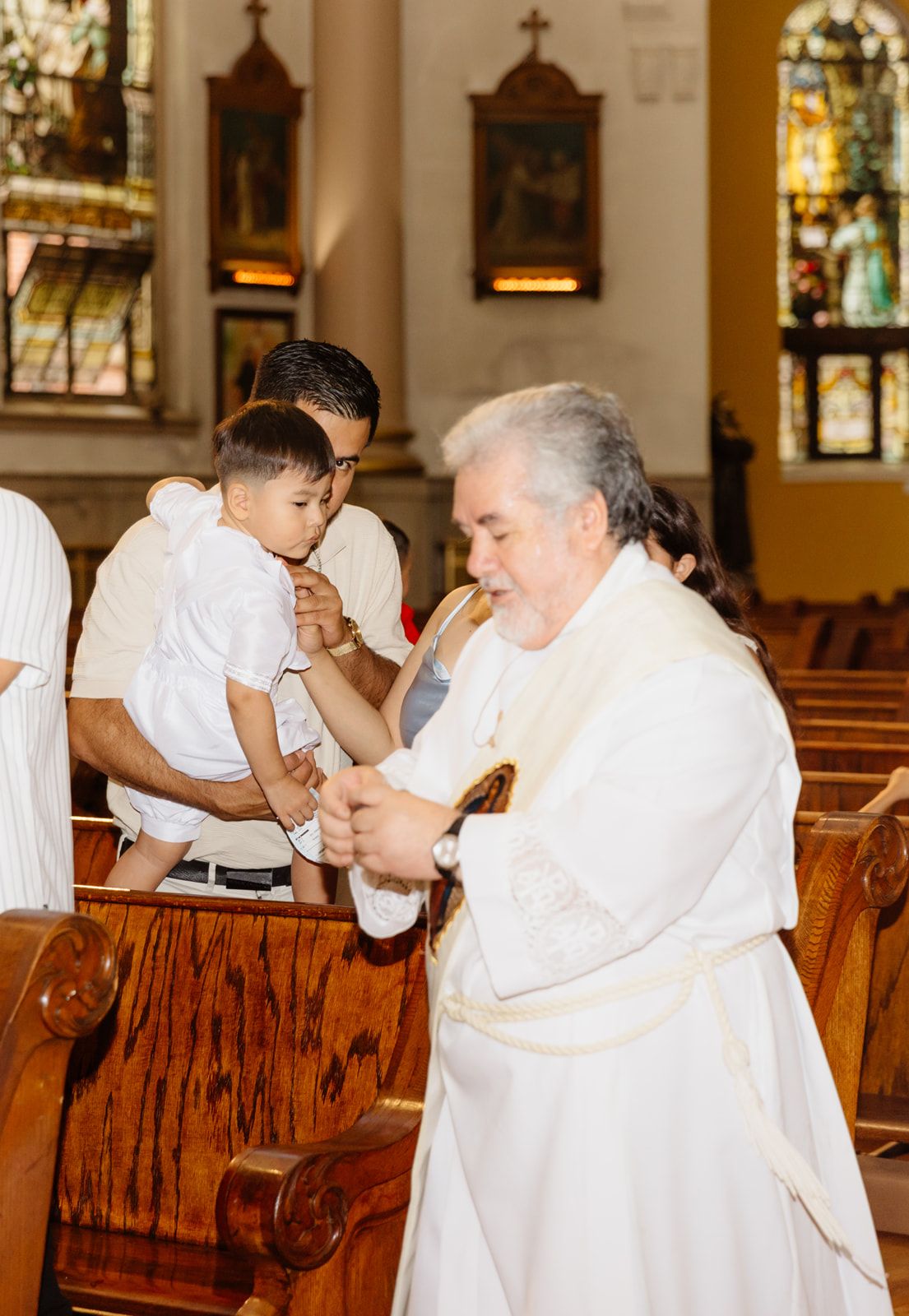 Priest in white robes interacts with a baby during a baptism ceremony inside a traditional Catholic church.