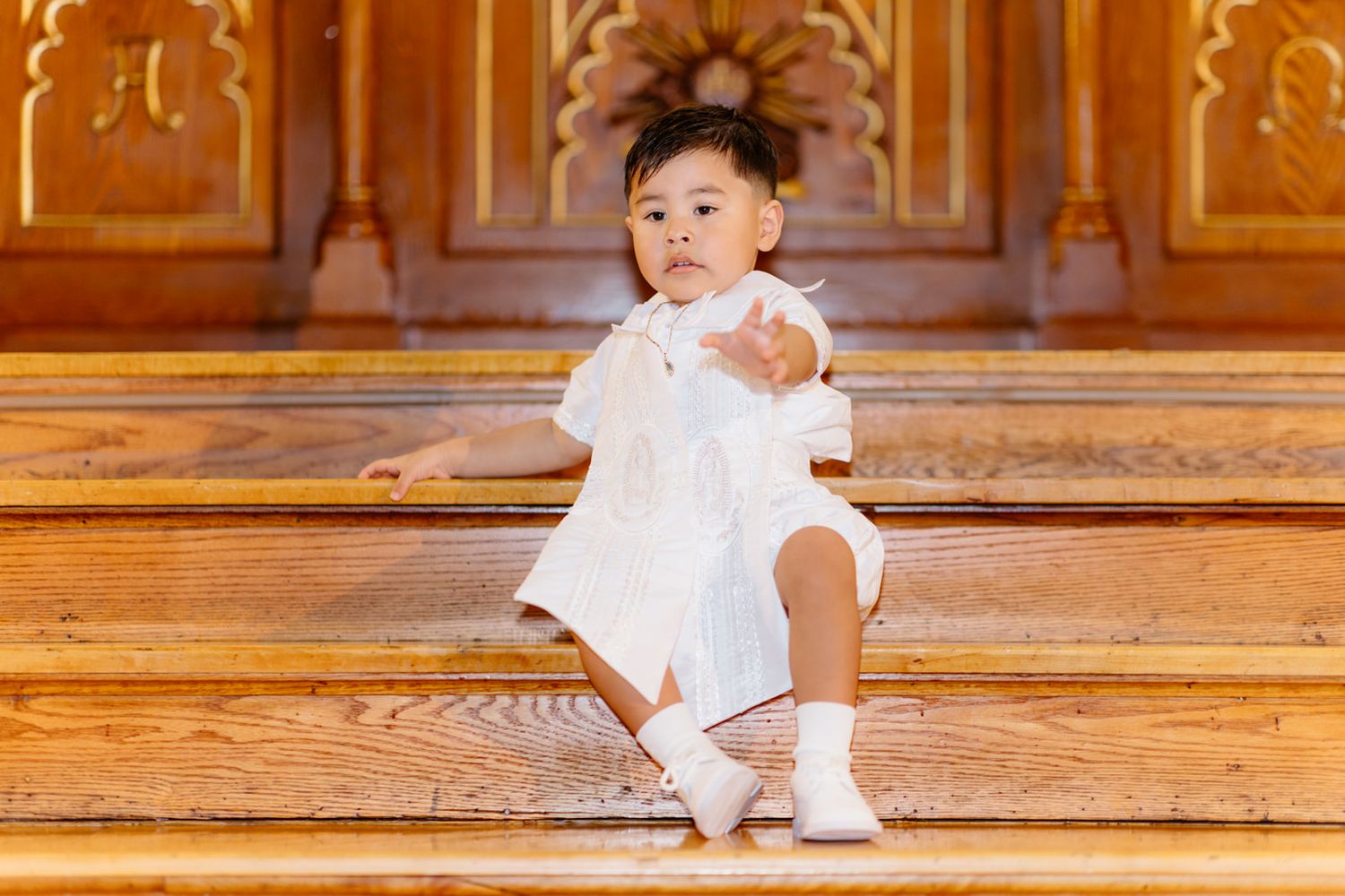 Baby in white christening outfit sits on wooden church steps reaching out with one hand.