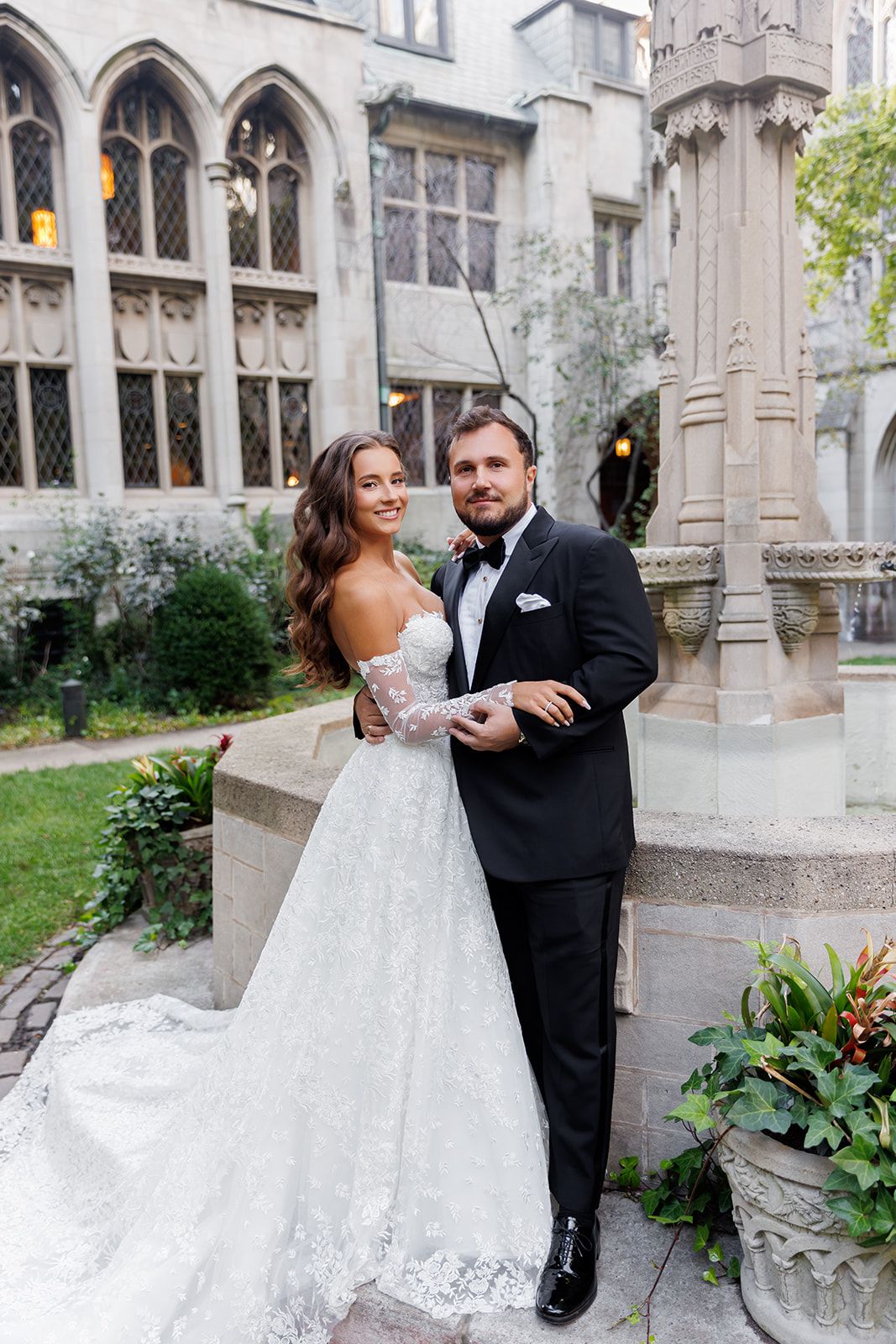 Bride in lace off-shoulder gown and groom in black tuxedo pose together outside a Gothic stone building.