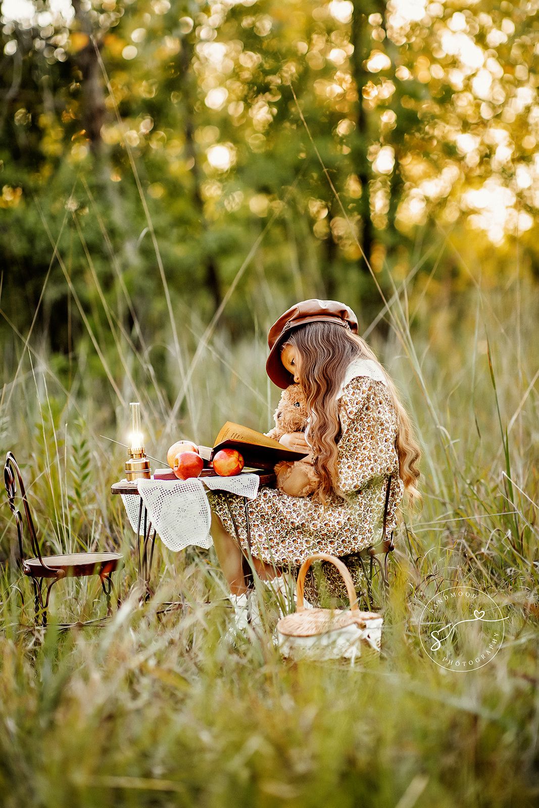 A young girl in a floral dress reads a book outdoors in tall grass with a teddy bear and small table nearby.