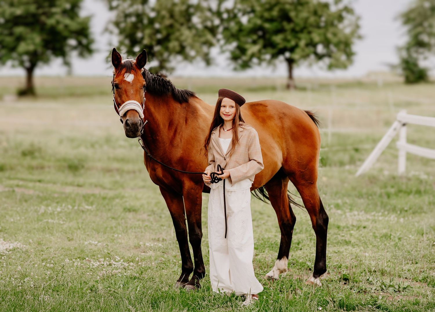 Young woman in beige outfit standing beside a bay horse in a lush green pasture with white fence.