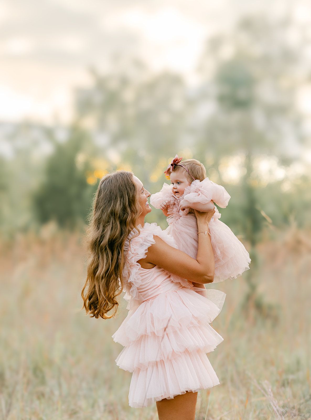Mother in pink tiered ruffle dress holding laughing baby girl in matching pink dress in meadow.