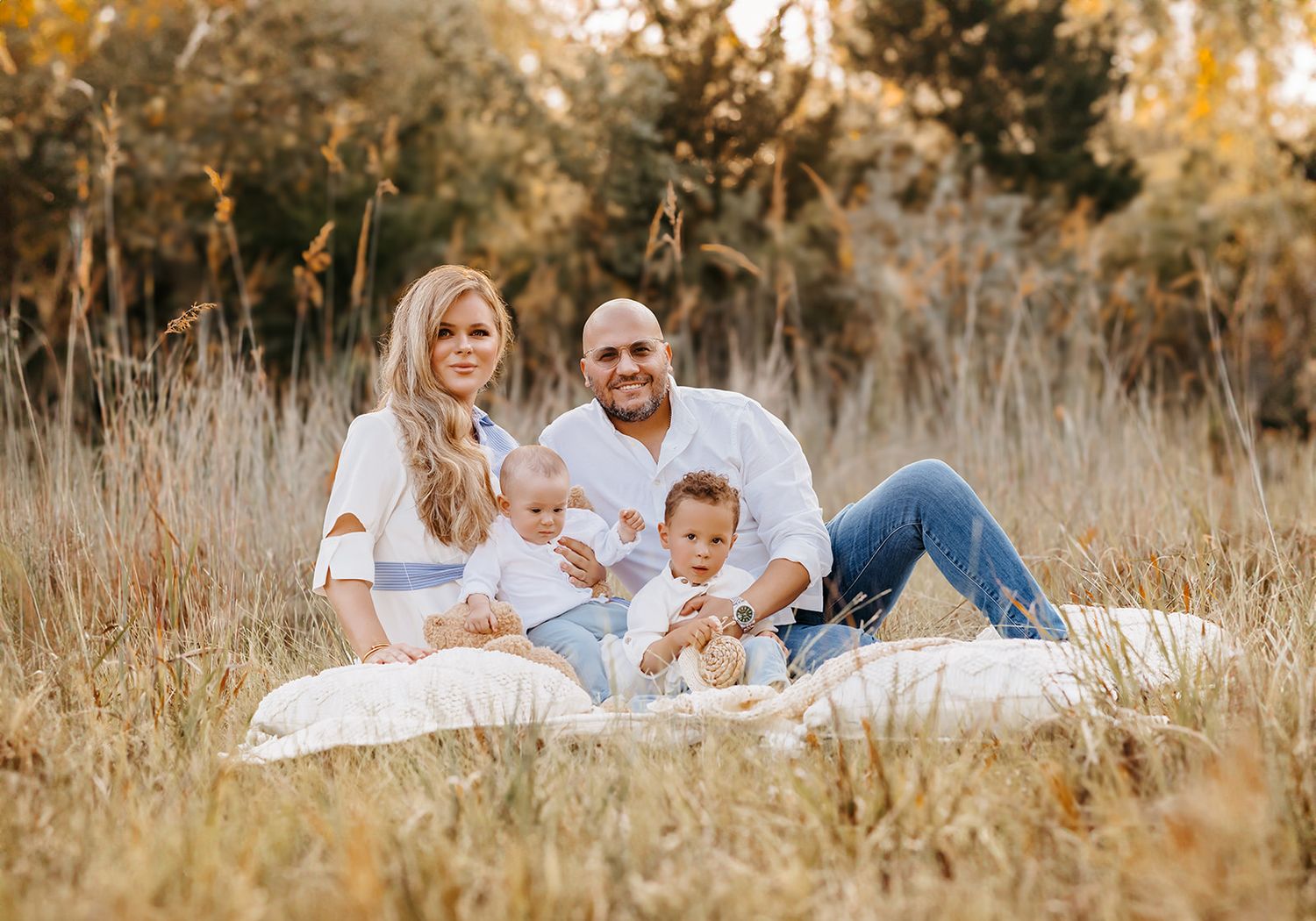 Family of four sitting on a white blanket in a golden field, parents smiling with two young toddlers on a sunny day.
