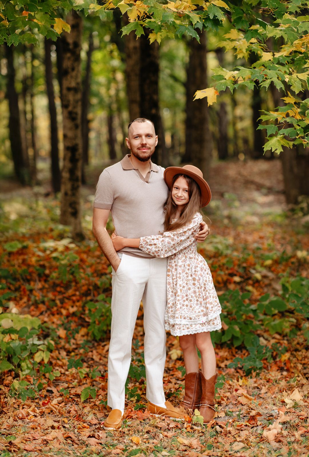 Couple embraces in autumn forest, woman in floral dress and brown hat, man in beige polo.