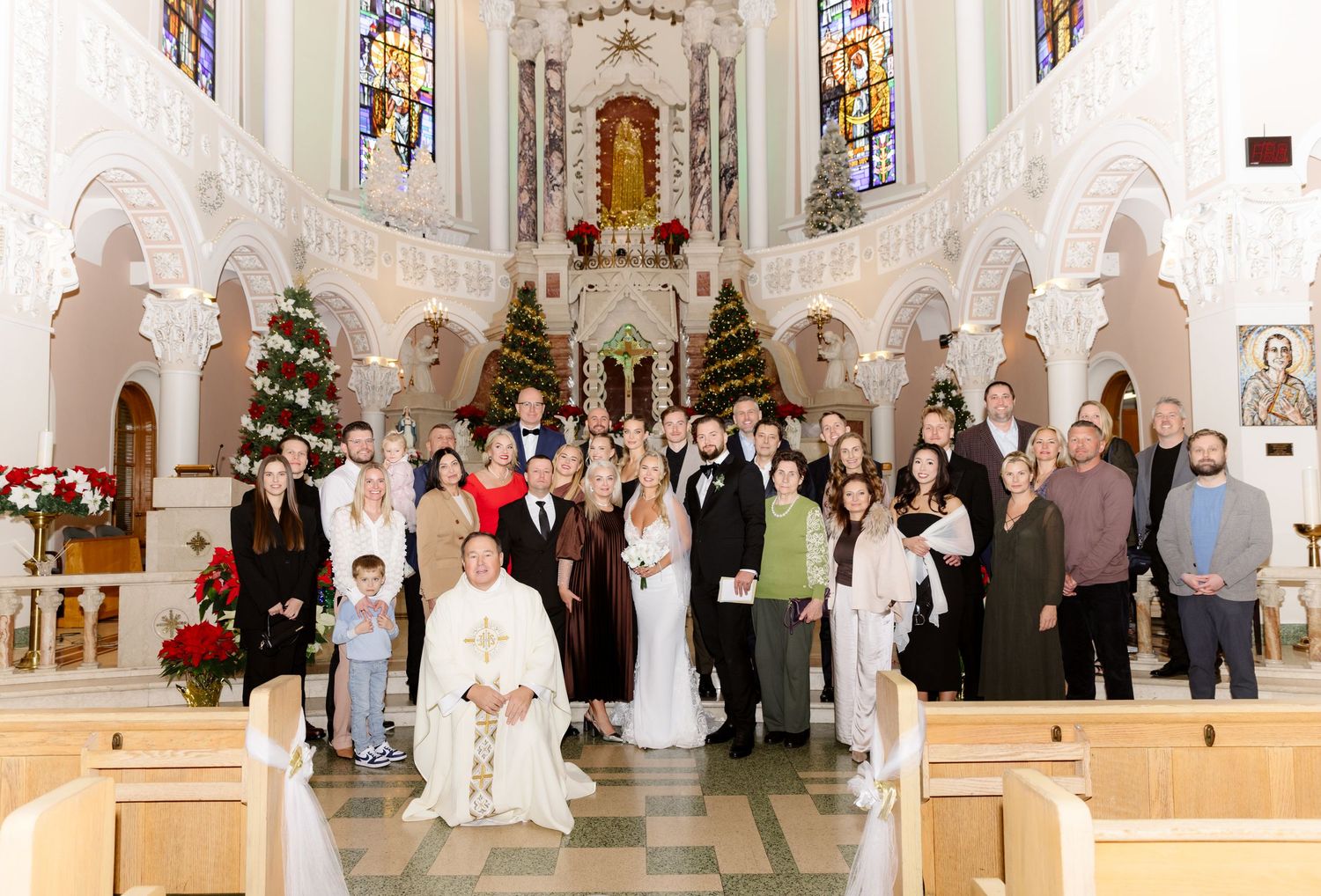 Wedding party and guests gather with kneeling priest for group portrait in Christmas-decorated Catholic church interior.