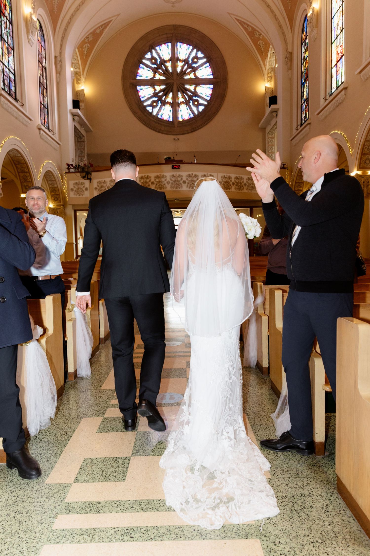Wedding guests applaud as bride in lace veil and groom walk down church aisle toward altar.