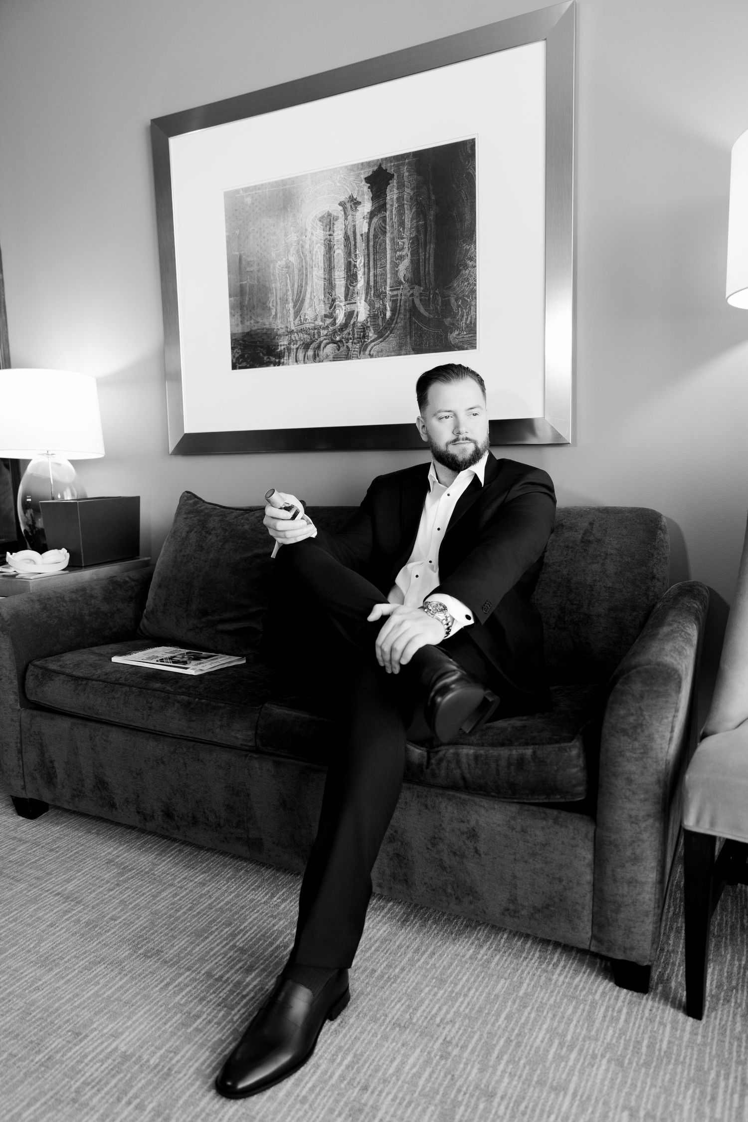 Elegant man in black suit sitting on velvet sofa holding drink in black and white portrait.