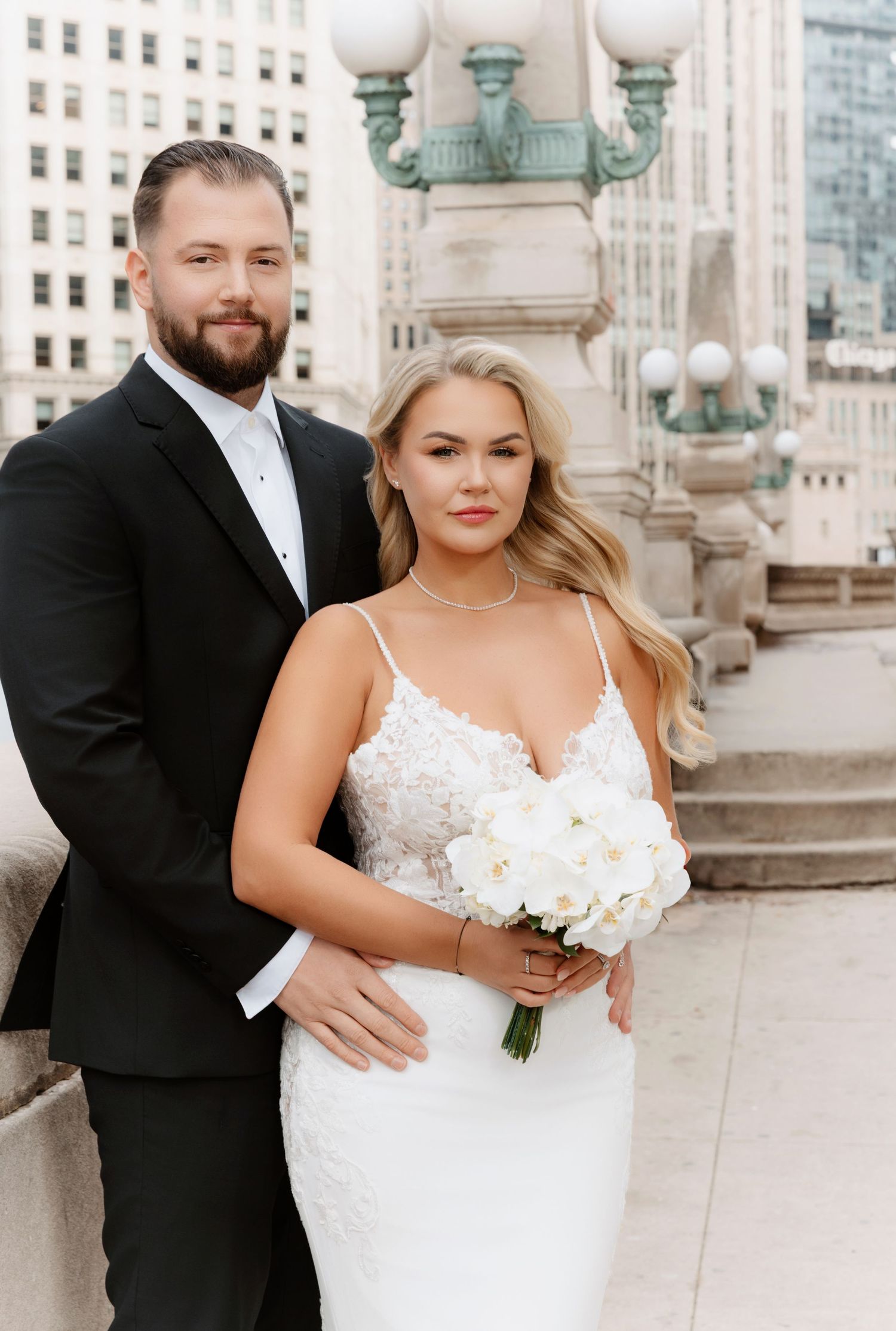 Bride in floral lace gown holds white orchid bouquet beside groom on ornate Chicago bridge.