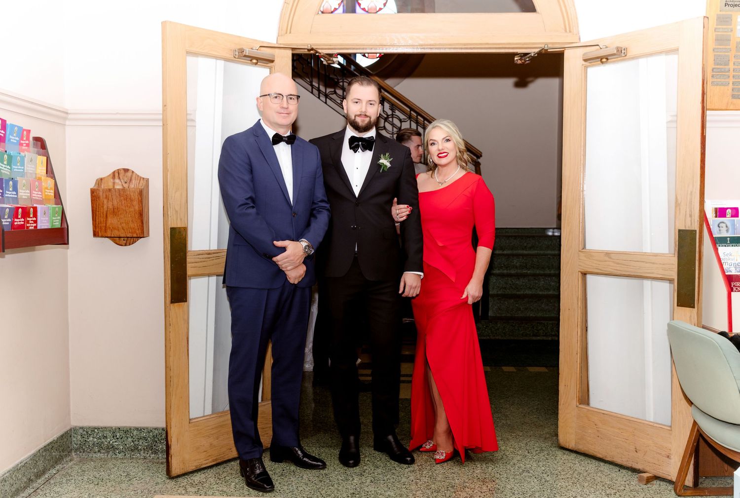 Groom flanked by father in blue tuxedo and mother in red dress posing at wedding venue doorway.