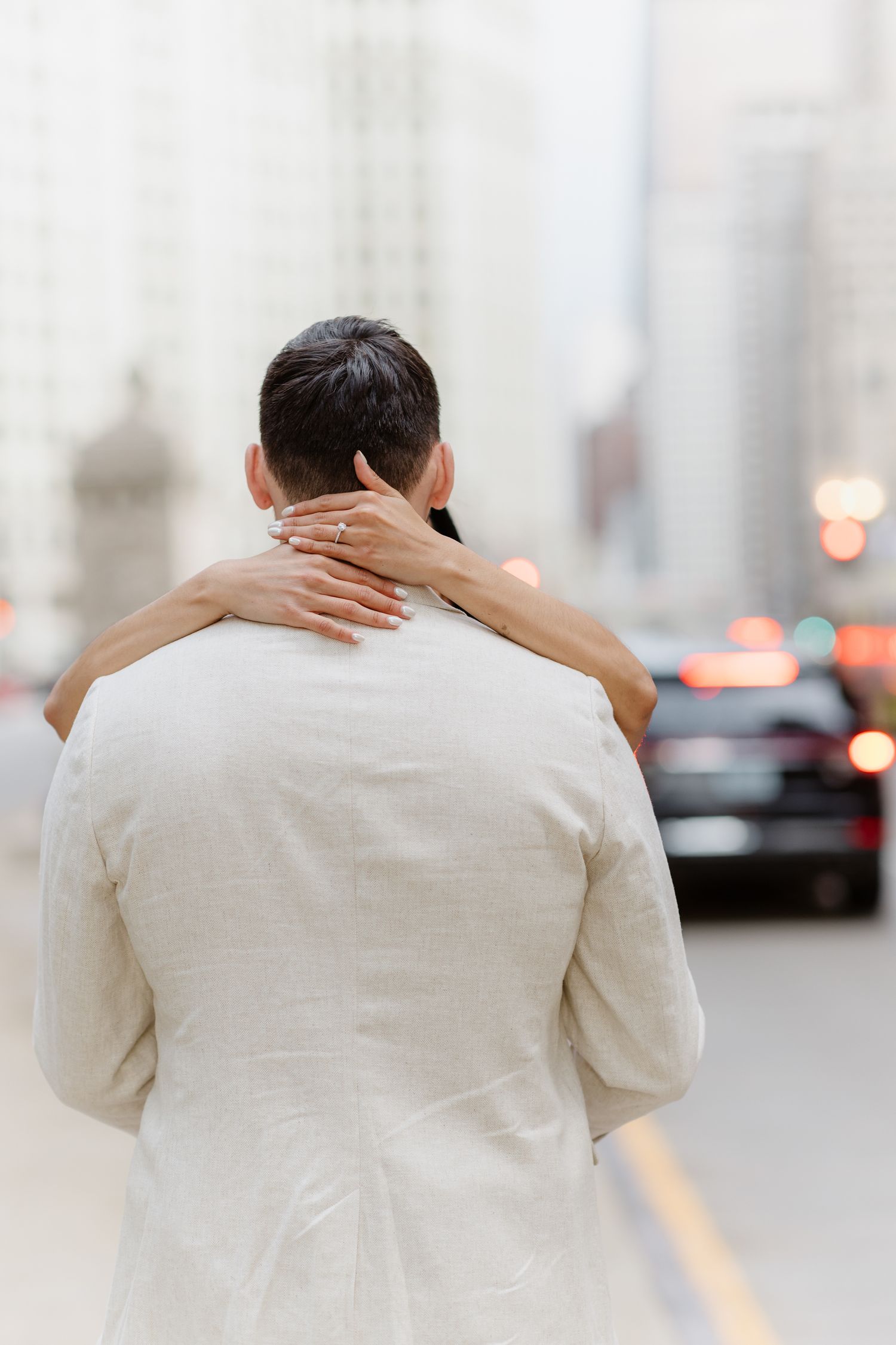 Couple embracing on a city street, woman's hand with engagement ring resting on man's shoulder.