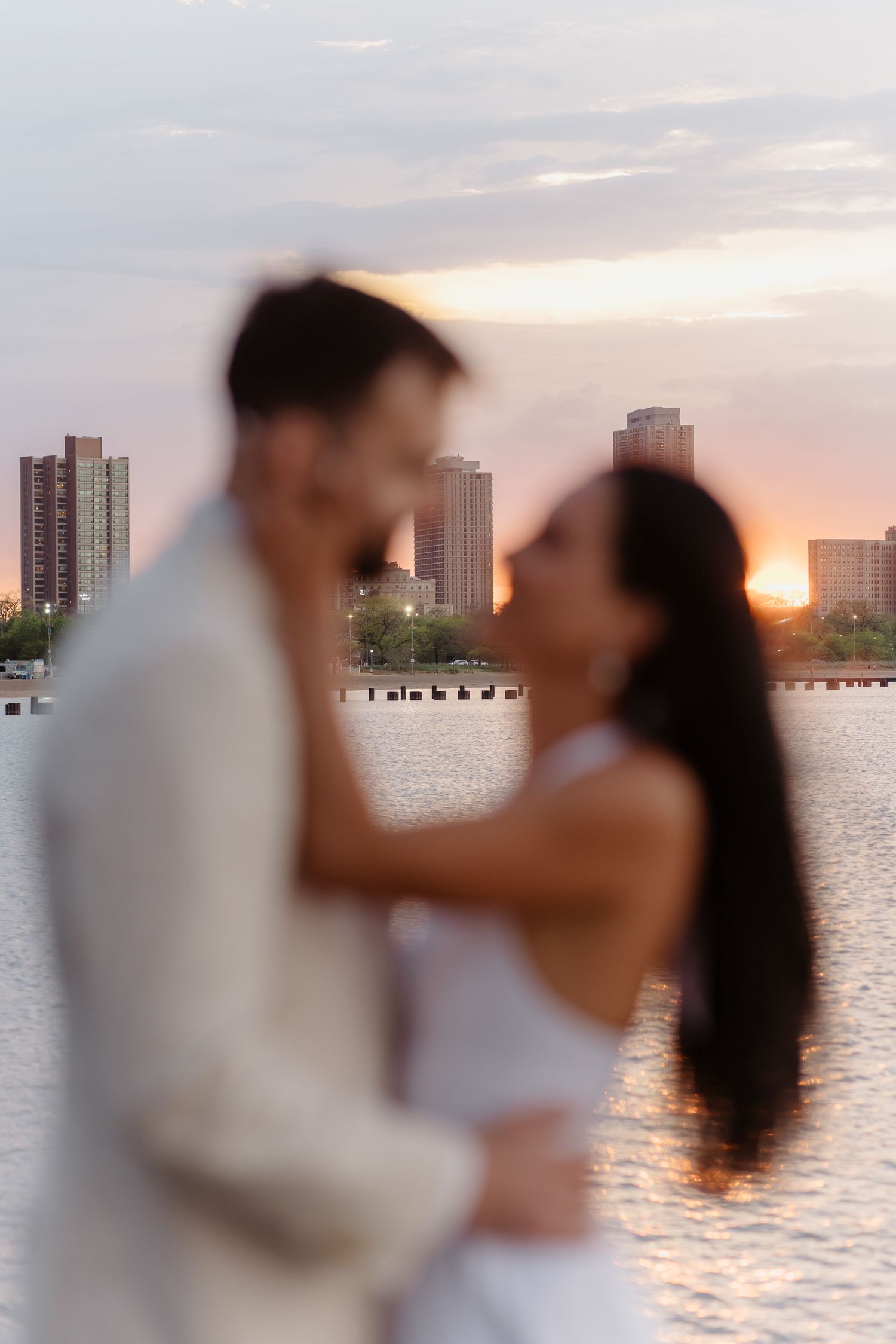 Couple sharing a kiss at sunset with city skyline reflecting on the water.