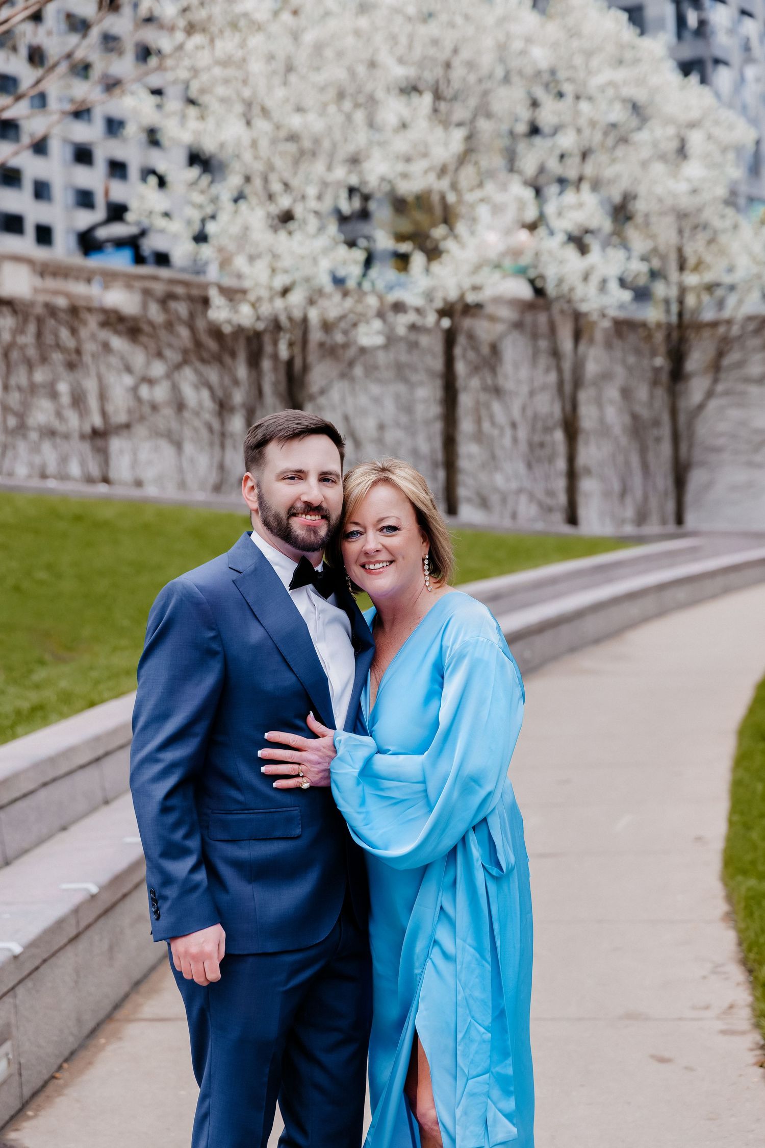 Groom in navy suit and woman in turquoise dress embrace near white blooming trees in urban park.