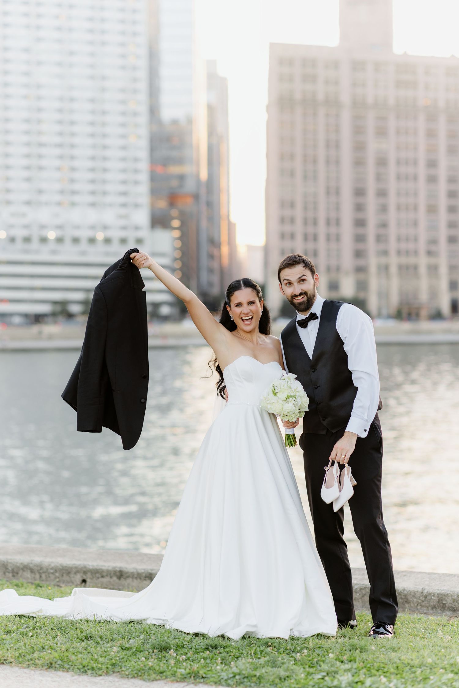 Joyful bride raises groom's jacket while he holds her shoes, posing by Chicago River at golden hour.