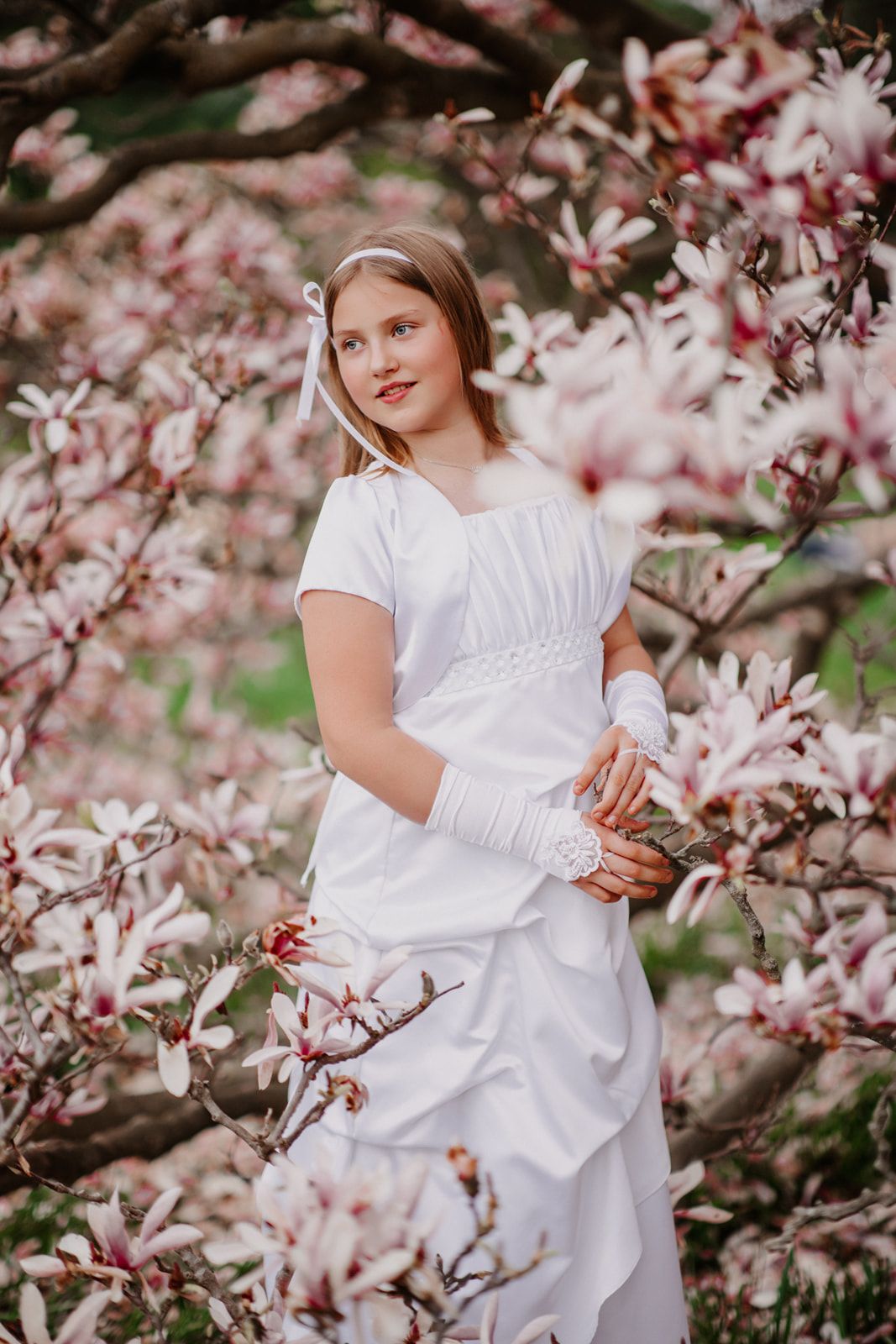 A young girl in a white First Communion dress and gloves poses gracefully among blooming pink magnolia trees.