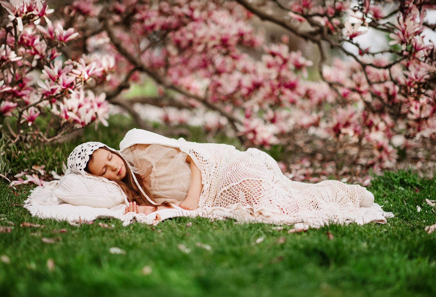 Baby sleeping peacefully outdoors on white blanket under blooming pink magnolia trees in spring garden.
