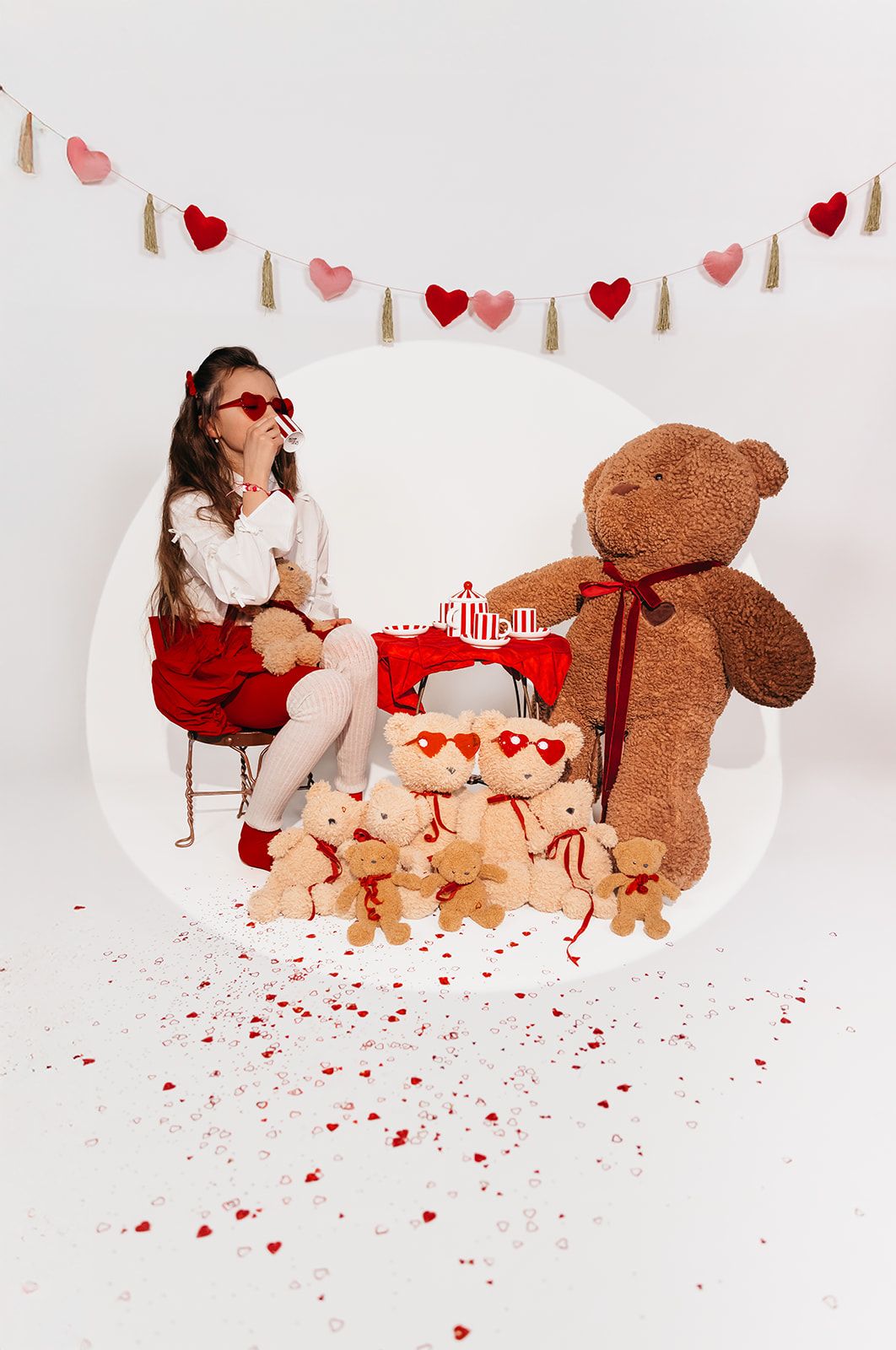 A girl in red skirt sits with teddy bears and Valentine decorations with hearts on white background.