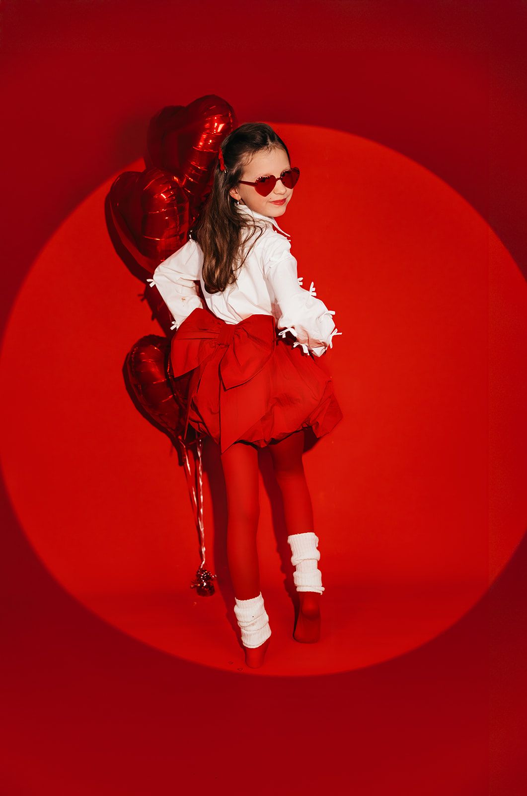 Young girl in red outfit with heart balloons and sunglasses poses against vibrant red background.
